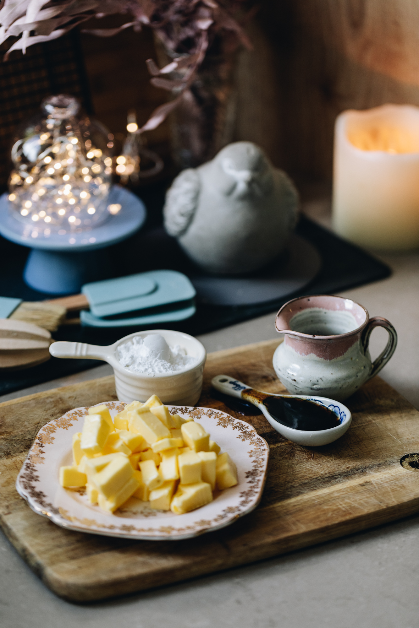 A wooden board sits on a brown stone bench. On the board is plate of cubed butter, a cup of icing sugar, a ceramic spoon of vanilla and a jug. Behind it blue and wooden spatulas, a wooden juicer, a pastry brush, a small blue cake stand, fairy lights inside a glass cover, pink dried flowers, a ceramic bird on a blue plate, a black cooling tray and a candle burning. 