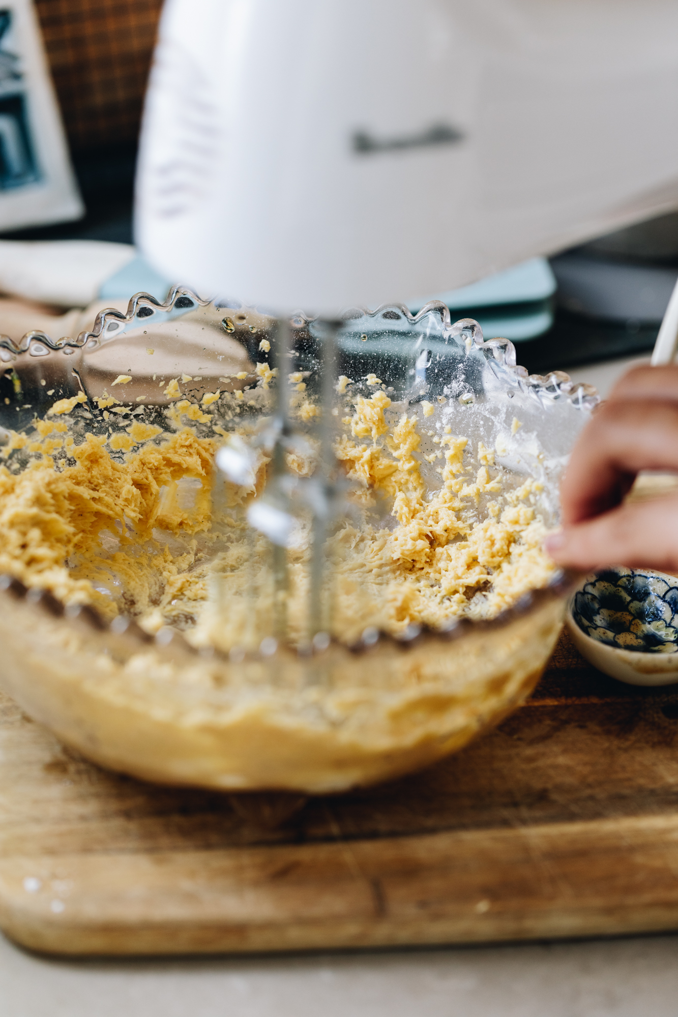 A glass vintage bowl sits on a wooden board, on brown stone bench. In the bowl is butter and sugar being whipped with a white hand mixer. There is baking utensils around the bowl and behind it. 