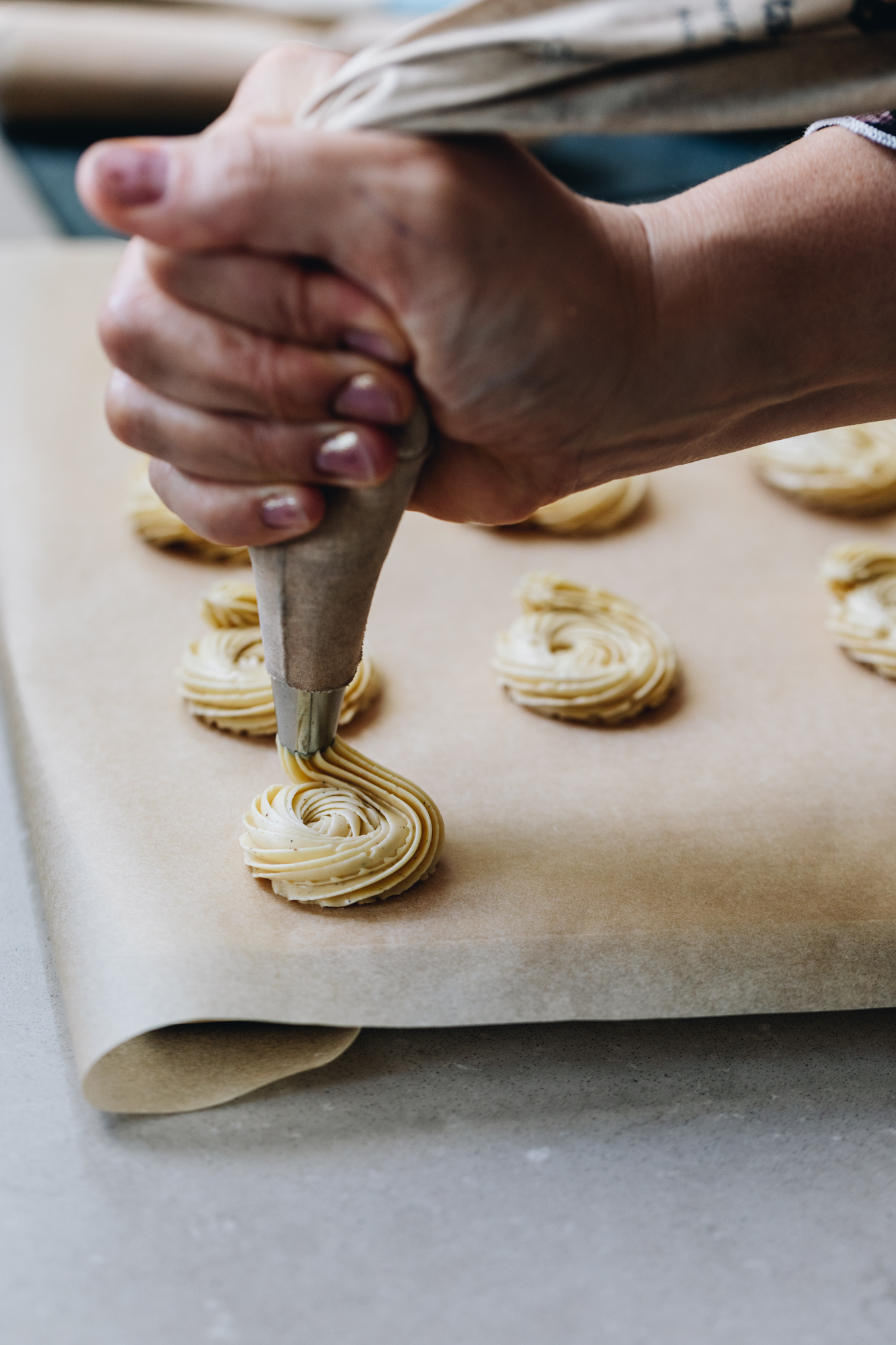 A wooden board is lined with brown baking paper and on the paper is round, piped rounds of the whipped vanilla butter recipe. Naomi is piping one with a white piping bag on the corner on the paper. 