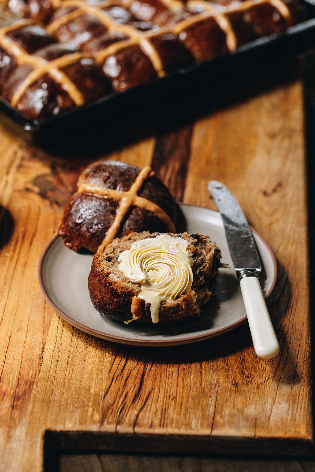 A wooden board sits on a wooden table. A brown ceramic plate is on the board and a hot cross buns is cut open with the cross part facing up. The other piece has the piped whipped vanilla butter recipe on it. It is melting gently down the front of the bun. A knife is also on the plate. A tray of hot cross buns sits behind the plate.