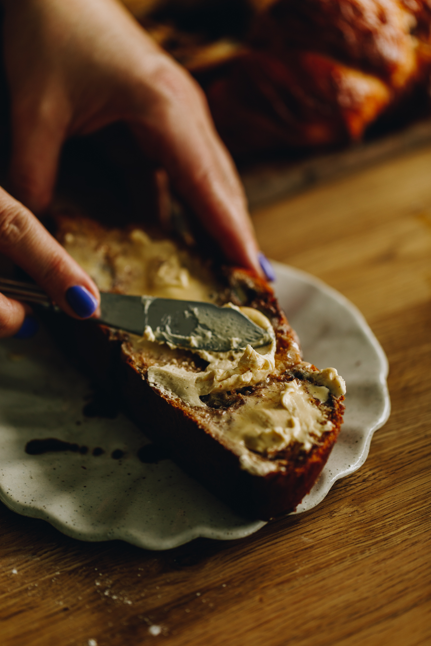 A crinkled ceramic plate sits on a wooden table. On the table is a piece of warm brioche bread that is being spread with a whipped vanilla butter recipe. Naomi is using a knife to spread it and it is starting to melt. 