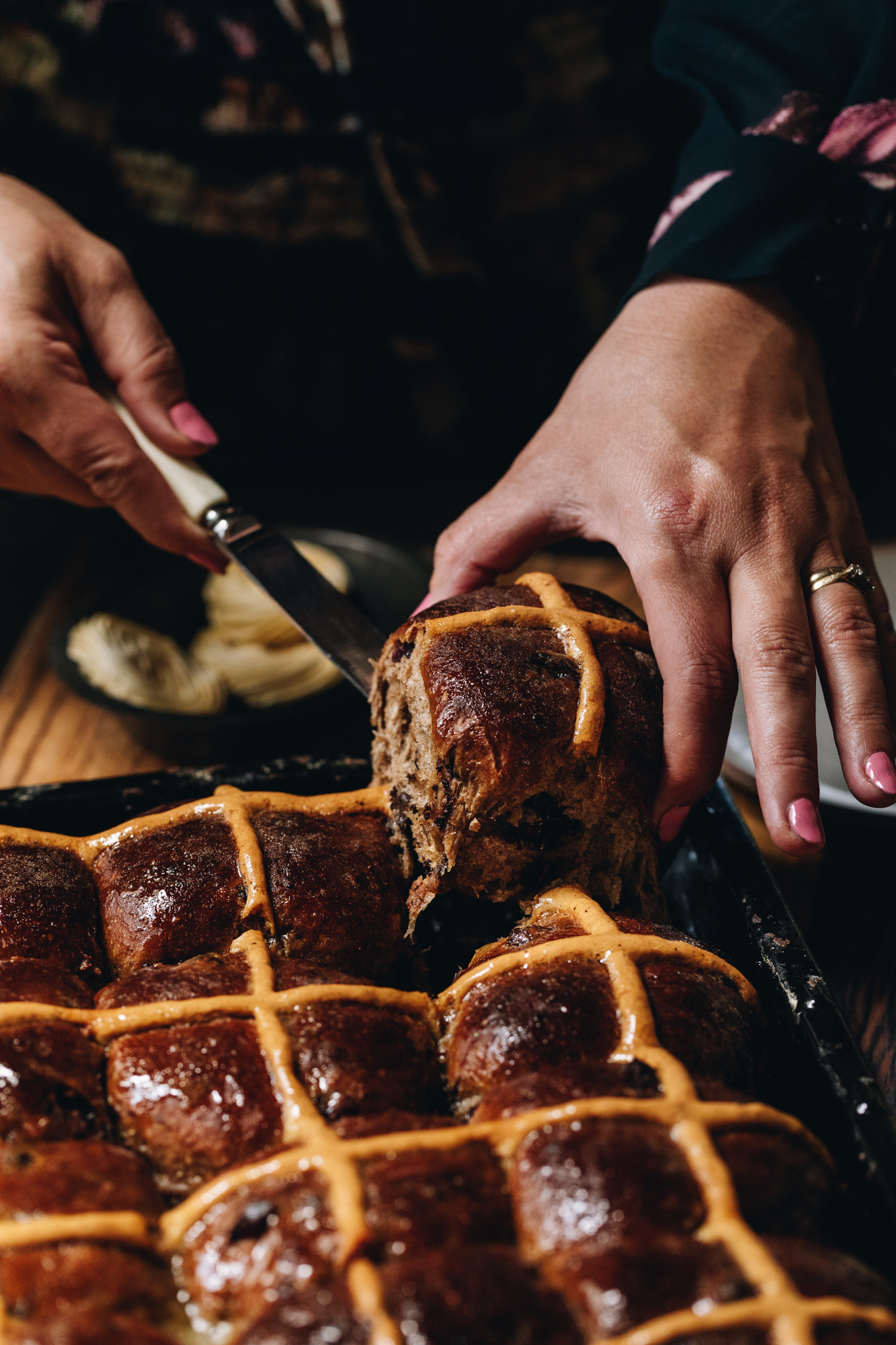A black tray with freshly baked and glazed spiced hot cross buns sits on a wooden table. Naomi is pulling on hot cross bun out of the tray with a bread and butter knife. A small plate of butter is behind the tray.