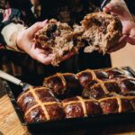 A tray of glazed overnight hot cross buns are in a black tray on top of a wooden table. Naomi has opened the buns showing the fluffy texture.