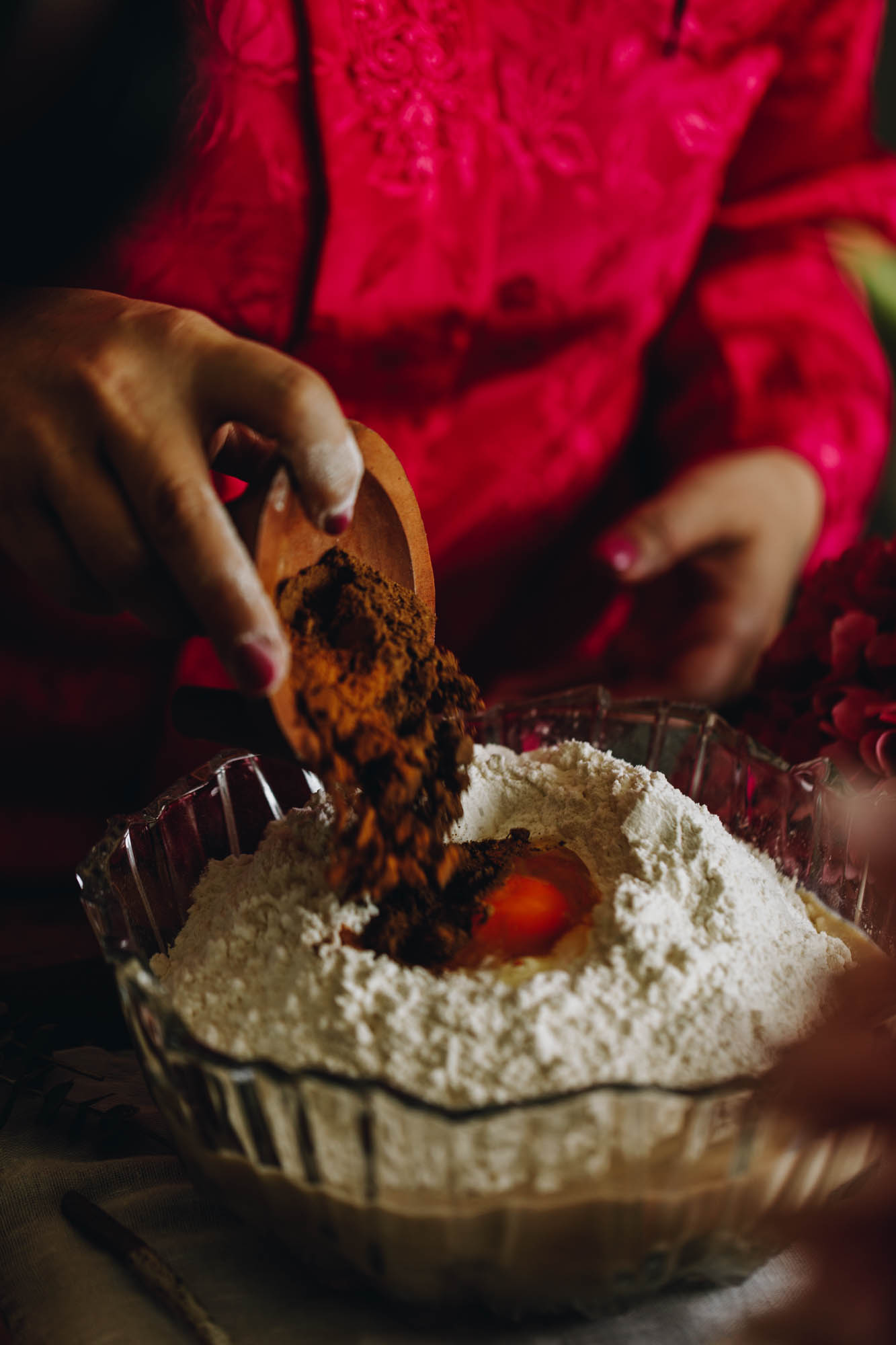 A vintage glass bowl sits on a wooden board. In the bowl is a flour mixture with an egg in the centre. Naomi is adding a small wooden container of spices in to it.