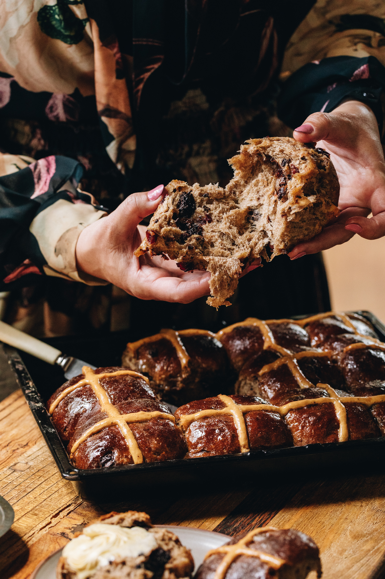 A tray of glazed spiced hot cross buns are in a black tray on top of a wooden table. Naomi has opened the buns showing the fluffy texture.