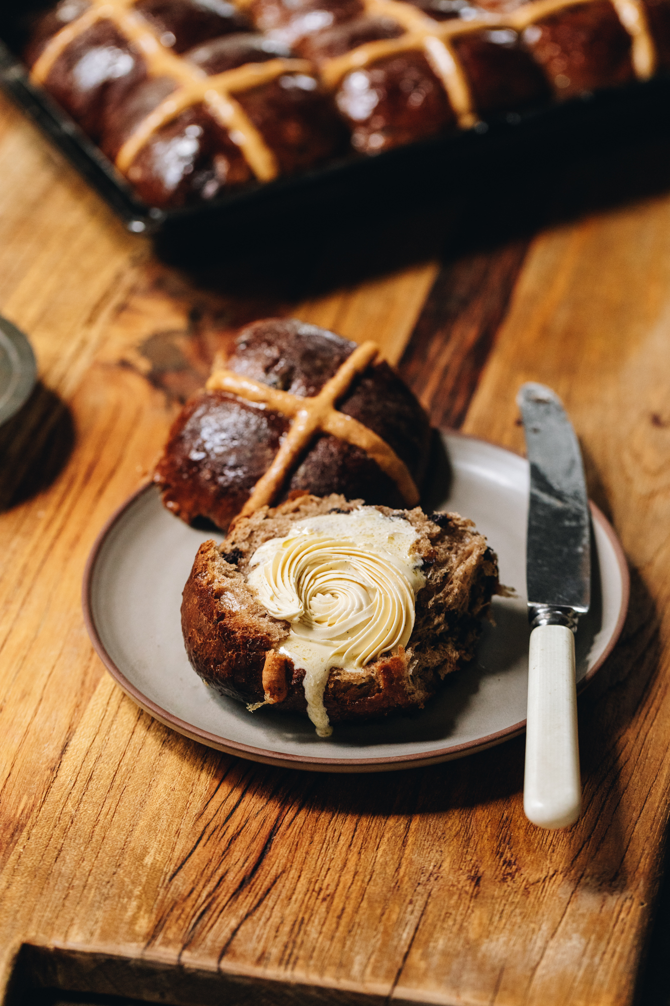 A wooden board sits on a wooden table. A brown ceramic plate is on the board and a spiced hot cross bun is cut open with the cross part facing up. The other piece has the piped whipped vanilla butter recipe on it that is drippy down the front of the bun. It is melting gently down the front of the bun. A knife is also on the plate. A tray of spiced hot cross buns sits behind the plate.