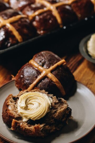 An overnight hot cross bun has been opened and is on a plate, it has piped butter on one side. Behind it is a tray of more hot cross buns.