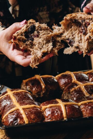 Spiced hot cross buns are freshly baked in a tray. One has been taken out by Naomi and she is breaking it open with her hands revealing the soft interior.