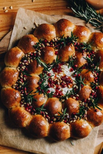 A whole baked brie cheese wreath is freshly baked and is sitting on a wooden board on baking paper. It is on a wooden table.