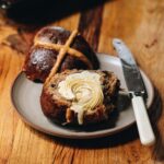 Whipped vanilla butter is melty down the front of a hot cross bun. It is starting to melt but the grooves of the piped butter are still seen.