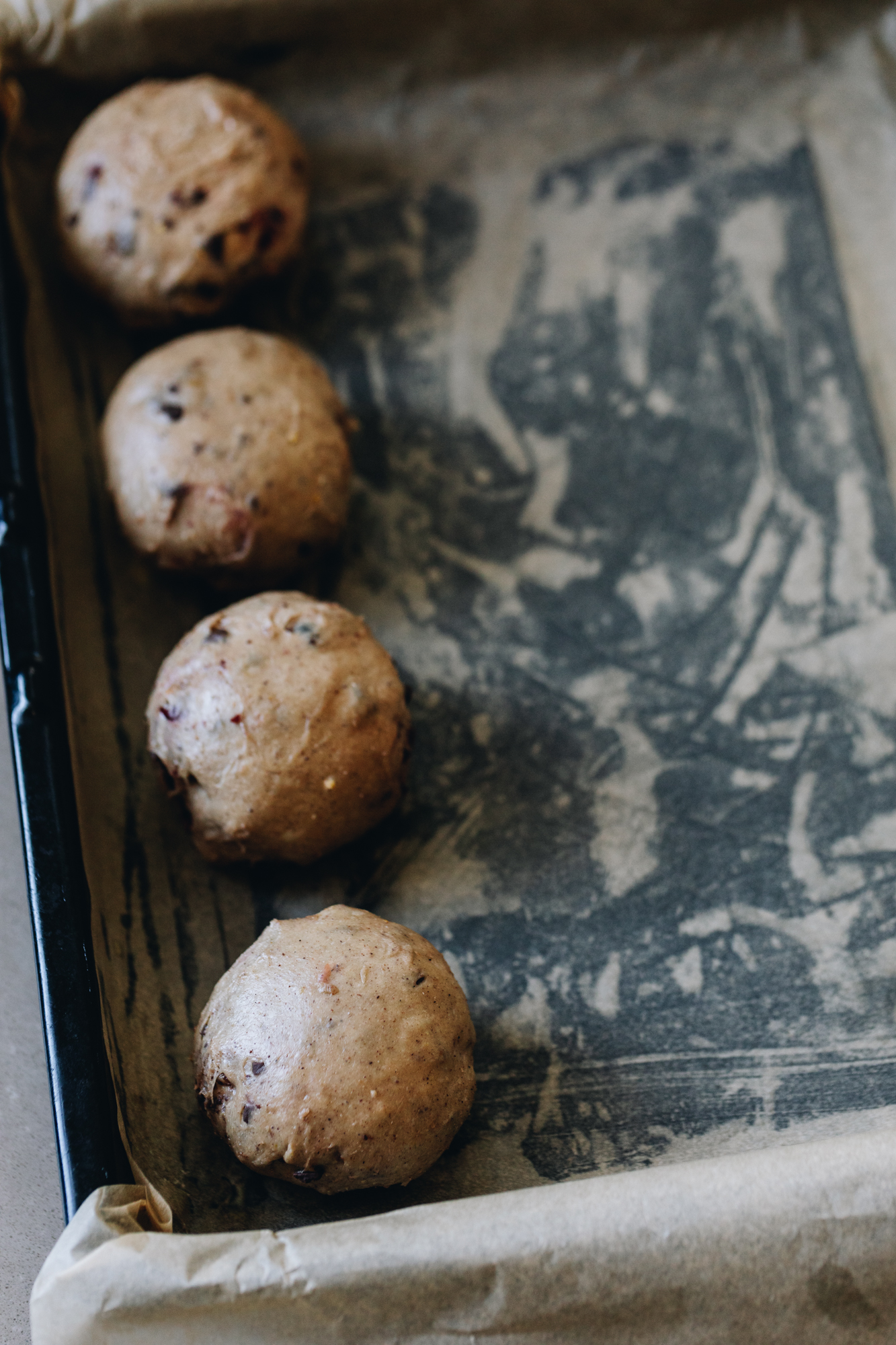A black tray is lined with brown baking paper and four spiced buns studded with dark chocolate are in a row in the tray. 