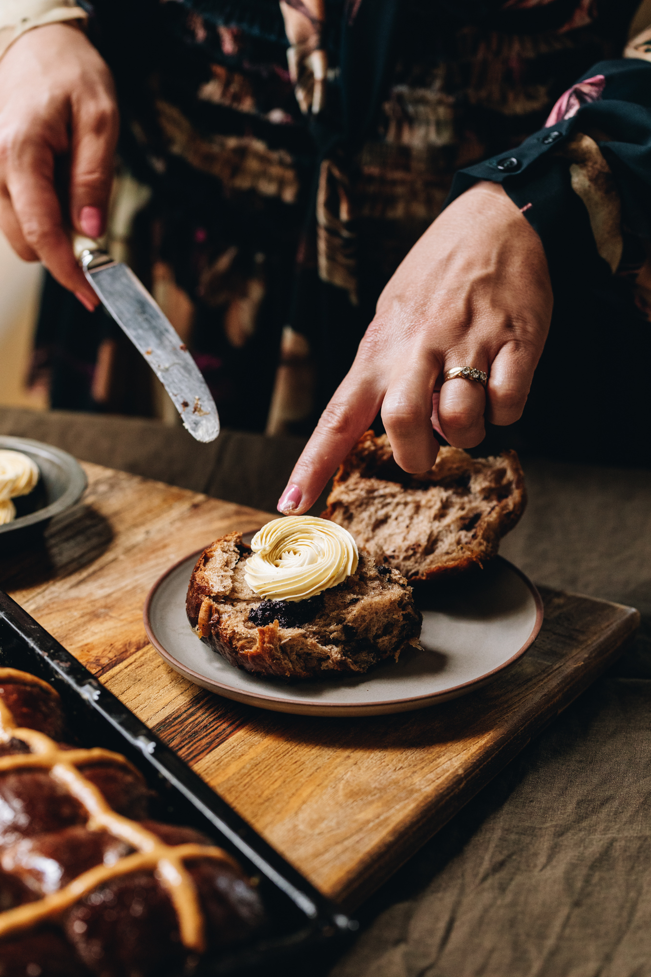 A wooden board sits on a wooden table. A brown ceramic plate is on the board and a overnight hot cross bun is cut open. One piece has the piped whipped vanilla butter recipe on it that Naomi has just placed down with a bread and butter knife. A tray of spiced hot cross buns sits in front of the plate.