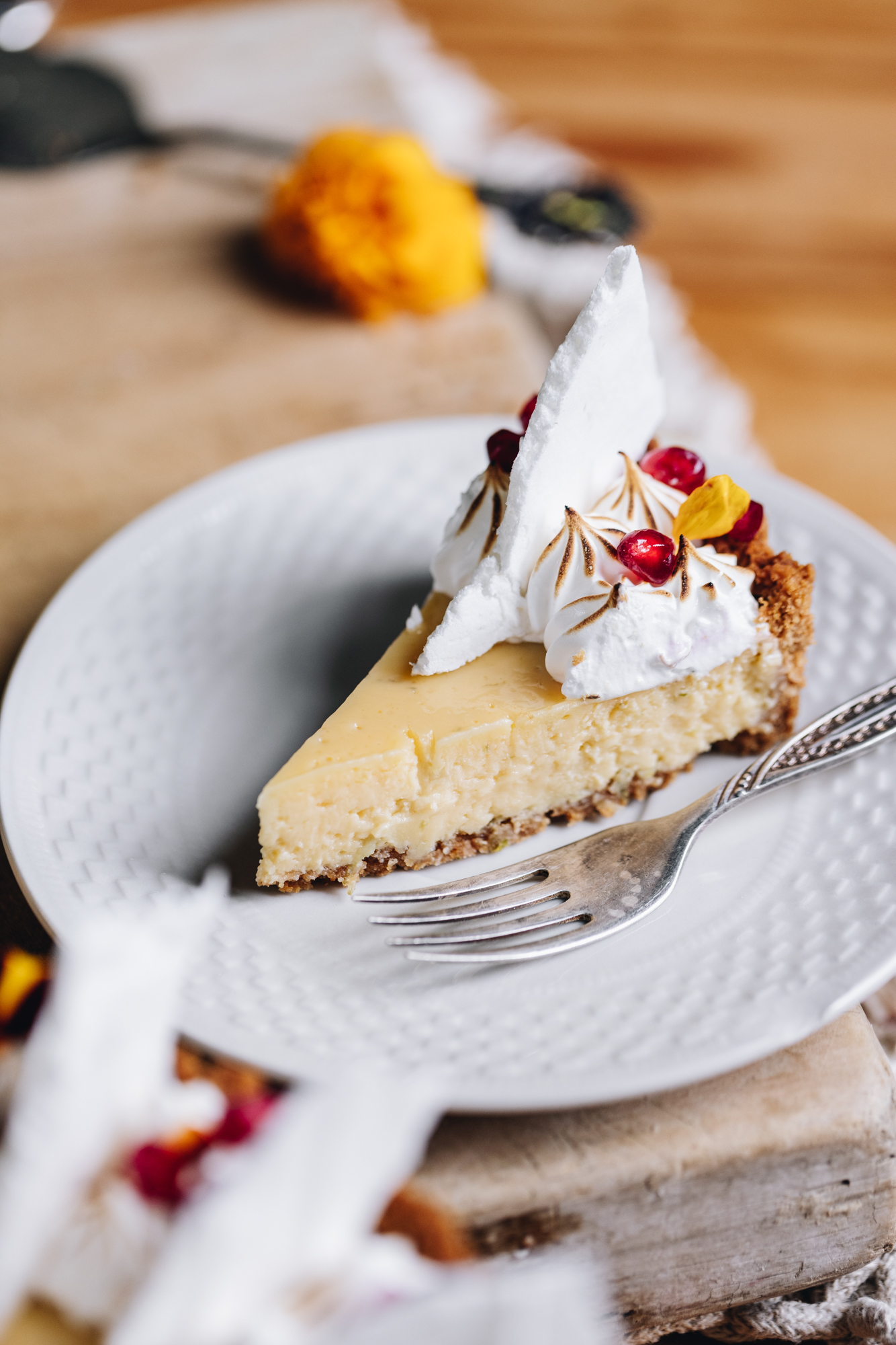 A piece of the key lime pie recipe is on a small white plate with a small fork next to it. It has been decorated with piped meringue and also has a couple of pomegranate seeds, an edible flower petal and a meringue shard on top of it. 