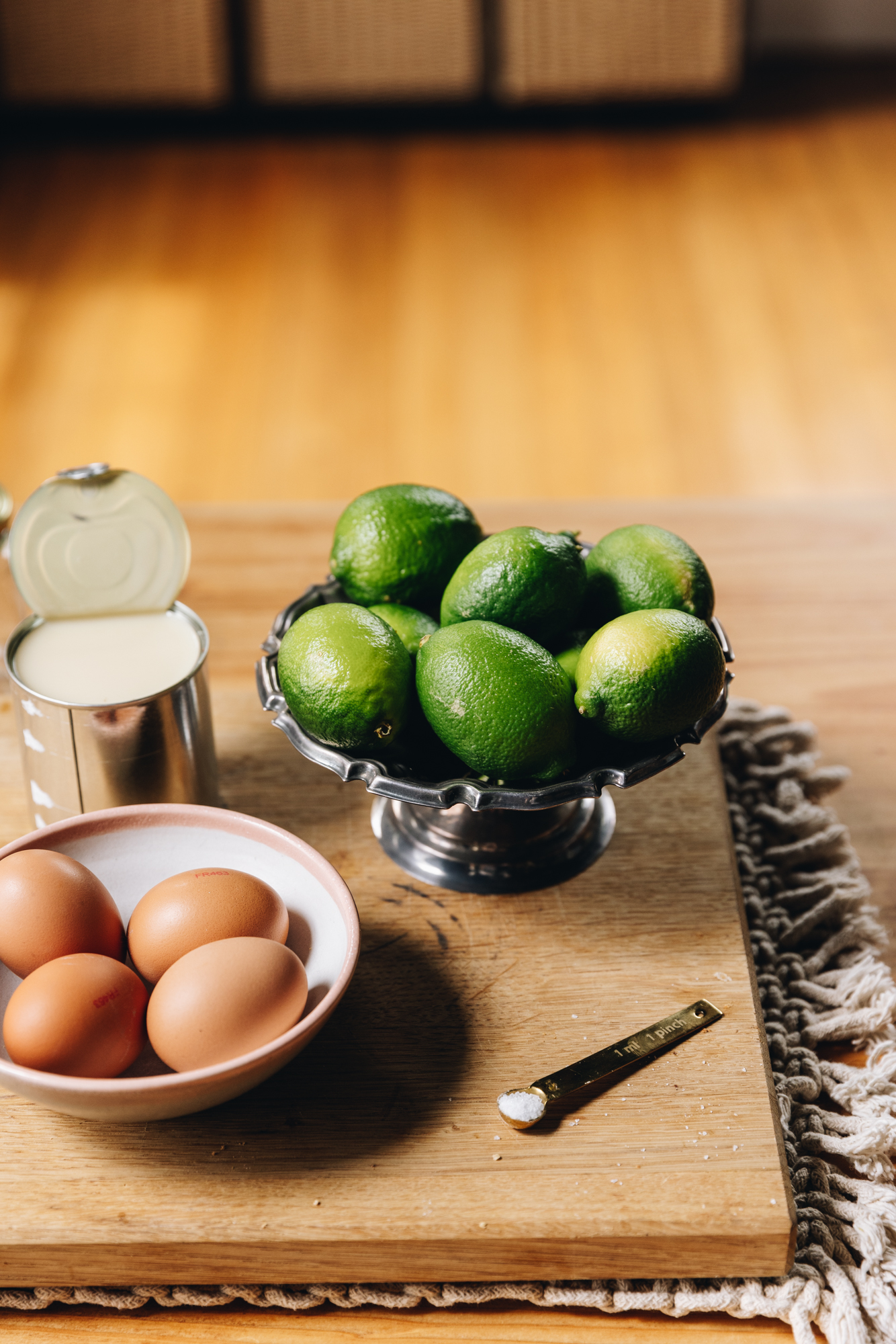 A wooden board sits on a weaved cream mat on top of a wooden table. On the board is a small bowl with eggs in it, a silver bowl with limes, a teaspoon of salt and an opened can of sweetened condensed milk. 