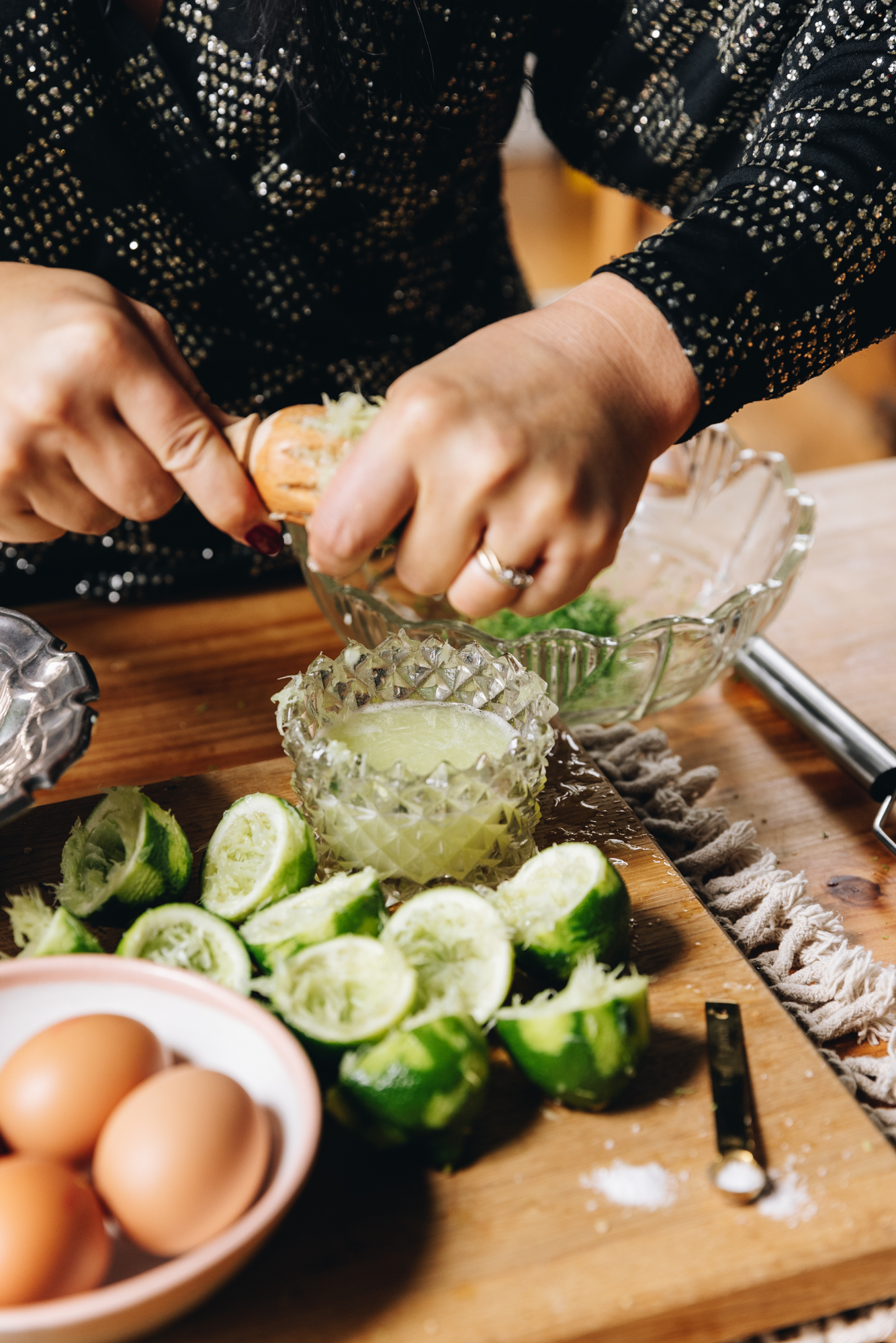 A wooden board sits on a weaved cream mat on top of a wooden table. On the wooden board at the front is a small bowl with eggs. Next to it is a pile of zest limes that have had the juice removed. A small vintage glass jar sits next to the limes and has lime juice in it. Naomi is using a wooden juicer to press the juice out of one lime. A glass vintage bowl is behind.