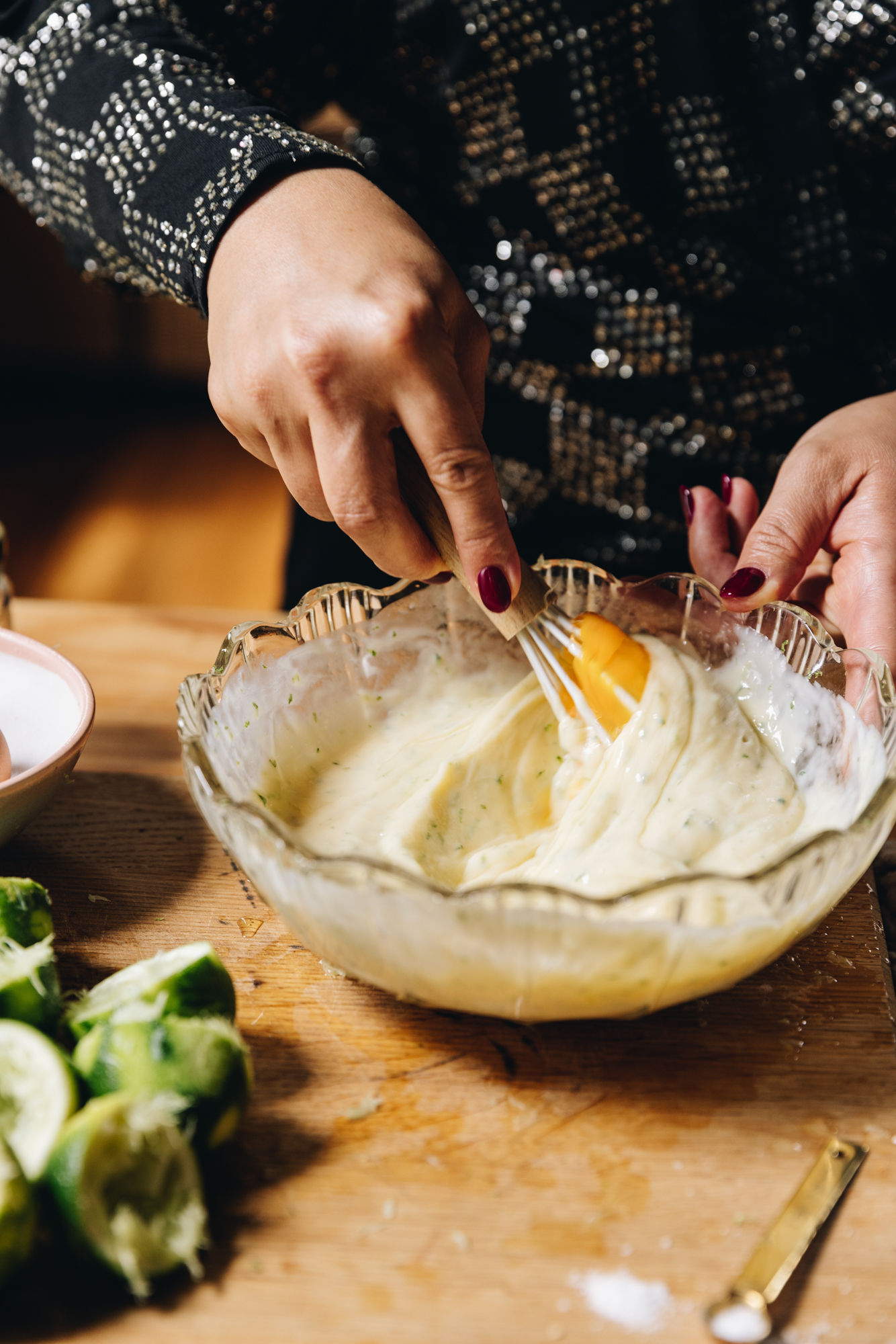 A wooden board sits on a wooden table. At the front is a pile of zested and juiced limes cut in half. Next to it is a glass vintage bowl that has a creamy lime mixture in it. Naomi is stirring it with a whisk and an egg yolk is visible. 
