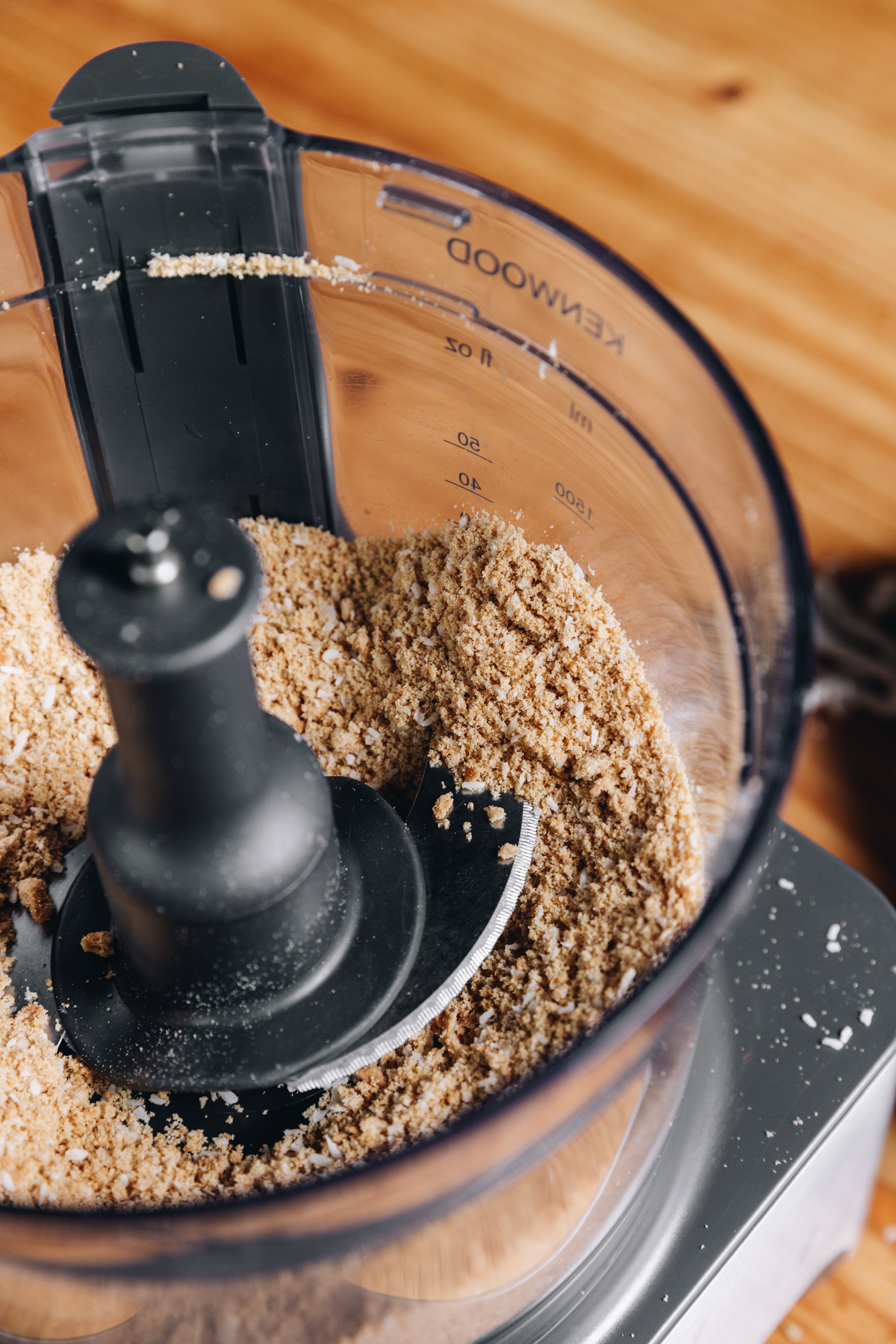 A food processor sits on a wooden table. The lid is off and in the processor is a biscuit and coconut crumb. 