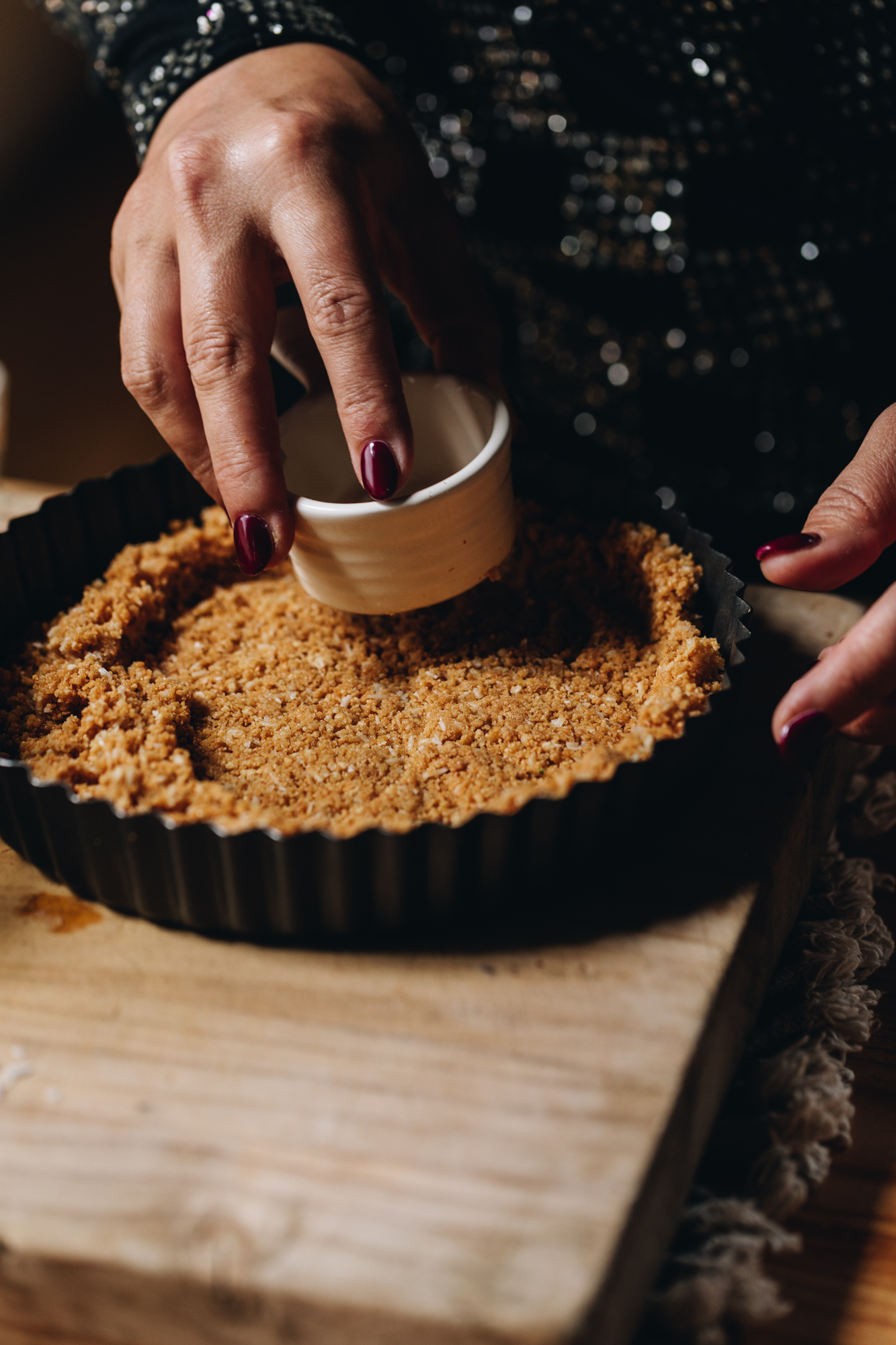 A wooden table sits on a weaved cream mat. On the board is a black quiche tin that has biscuit crumbs in it. Naomi is using a white cup to press the crumb in to the tin. 
