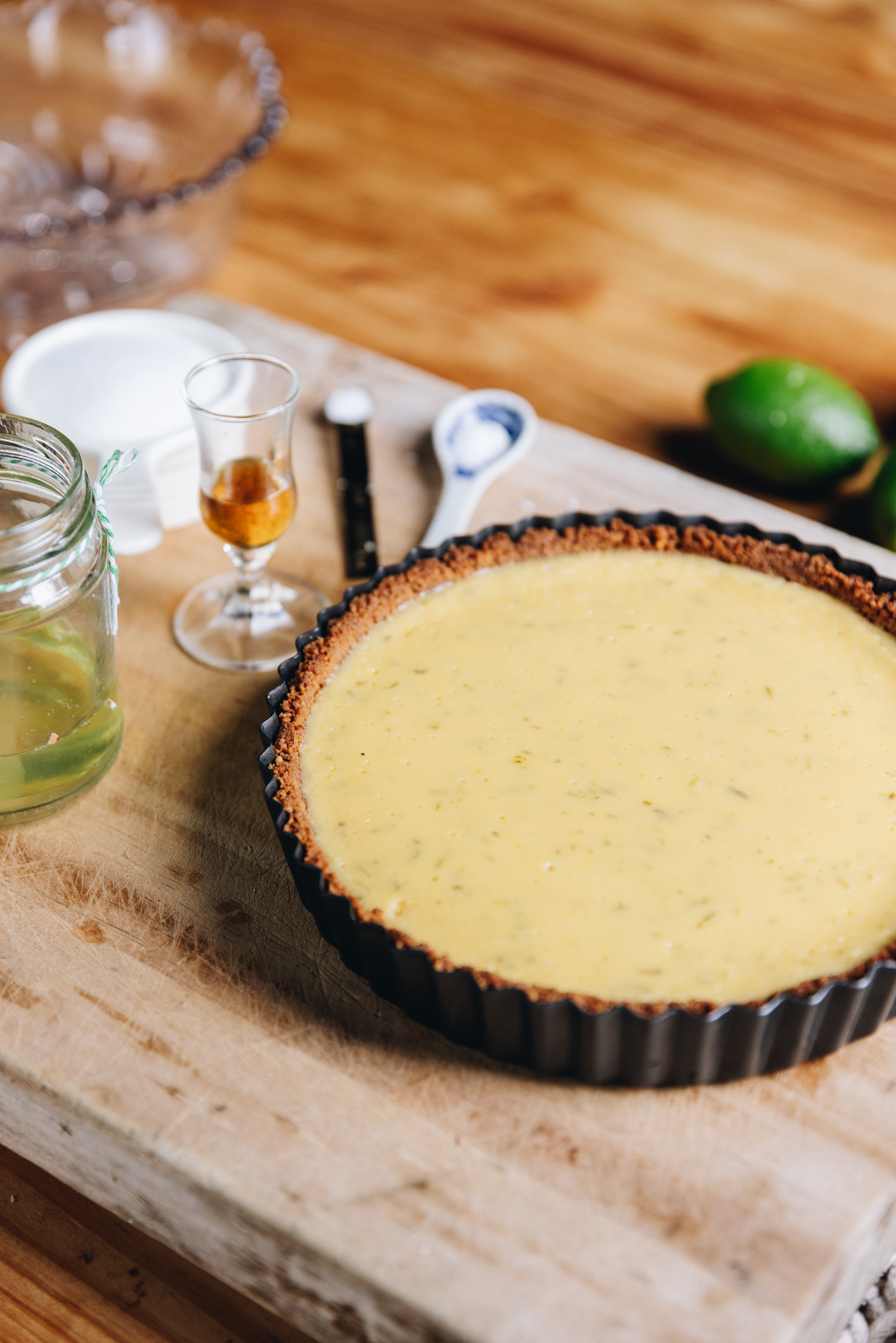 A wooden board sits on a wooden table. On the board is a freshly baked key lime pie recipe that has been baked in a black quiche tin. Next to it is a little jar of vanilla and egg whites along with some spoons of salt and cream of tartar. A lime is on the table next to it. A vintage glass bow is in the background.  