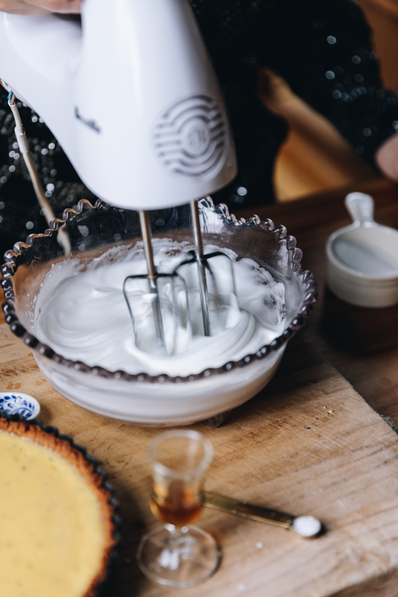 A wooden board is on a wooden table. To the front of the photo is a baked key lime pie. A small glass of vanilla and a spoon of cream of tartar sit next to it. A glass vintage bowl is also on the board, it has the fresh meringue recipe in it and it is being whipped with a white hand mixer. A cup of sugar is next to it.