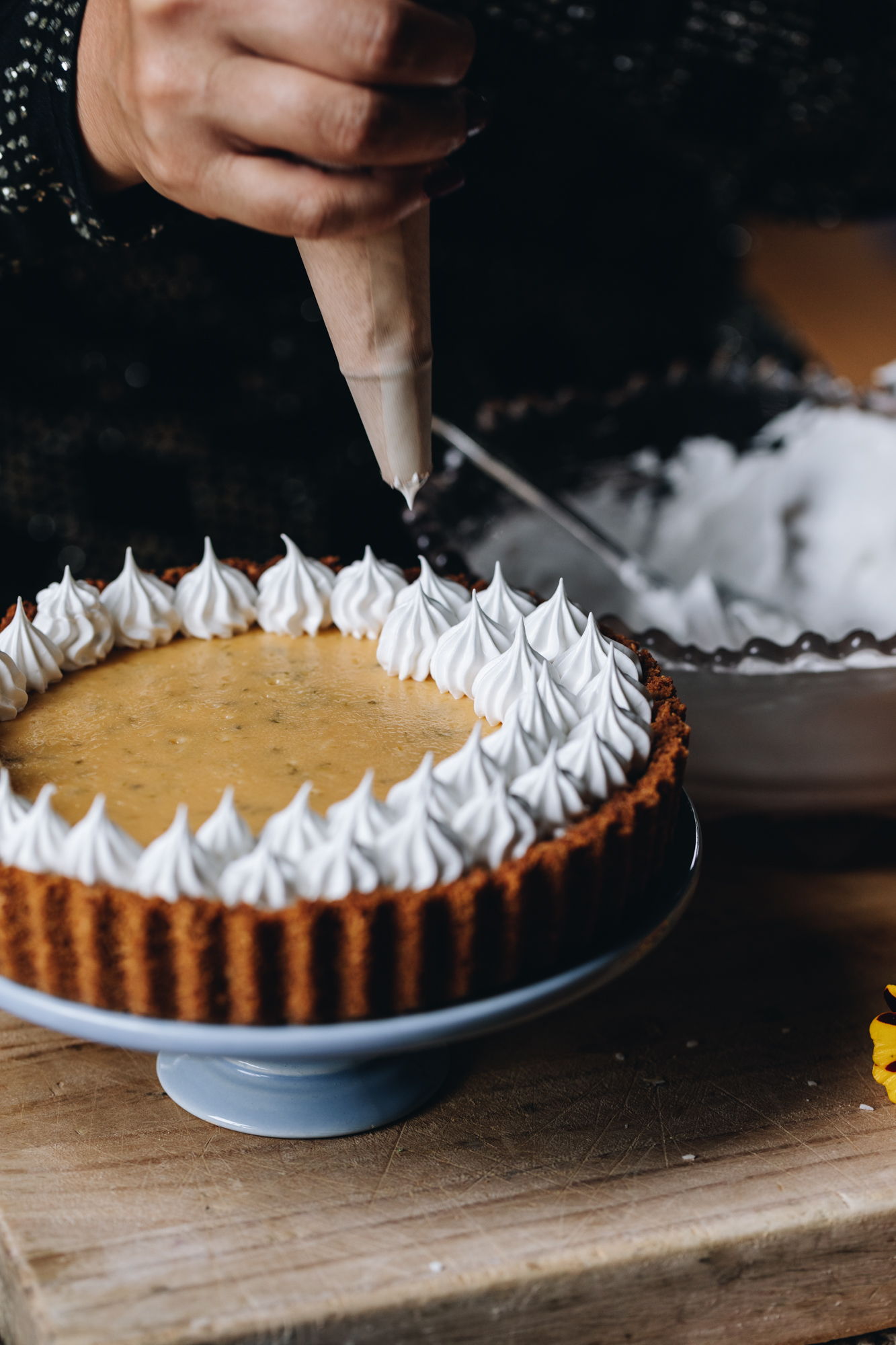 A key lime pie recipe is on a small blue cake stand on a wooden board. A bowl with meringue is sitting behind it. Naomi is piping a French meringue recipe on to the pie using a piping bag. There is two rows of piped meringue.