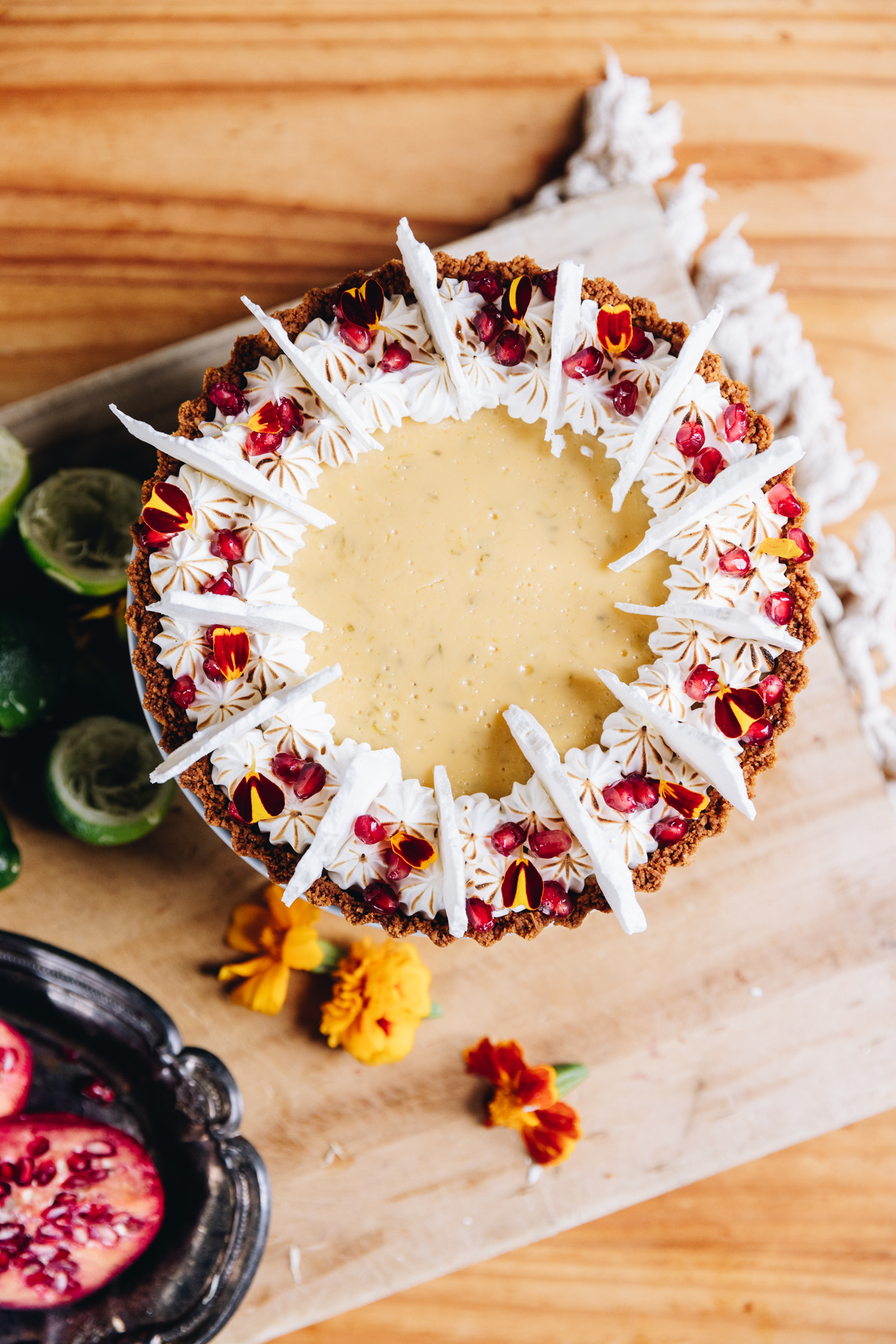 A flat lay shot shows a freshly decorated key lime pie recipe that has been decorated with piped meringue, meringue shards, pomegranate seeds and flower petals. The pie is on a wooden board and around the pie is cut limes, pomegranate pieces in a black small tray and some orange edible flowers.