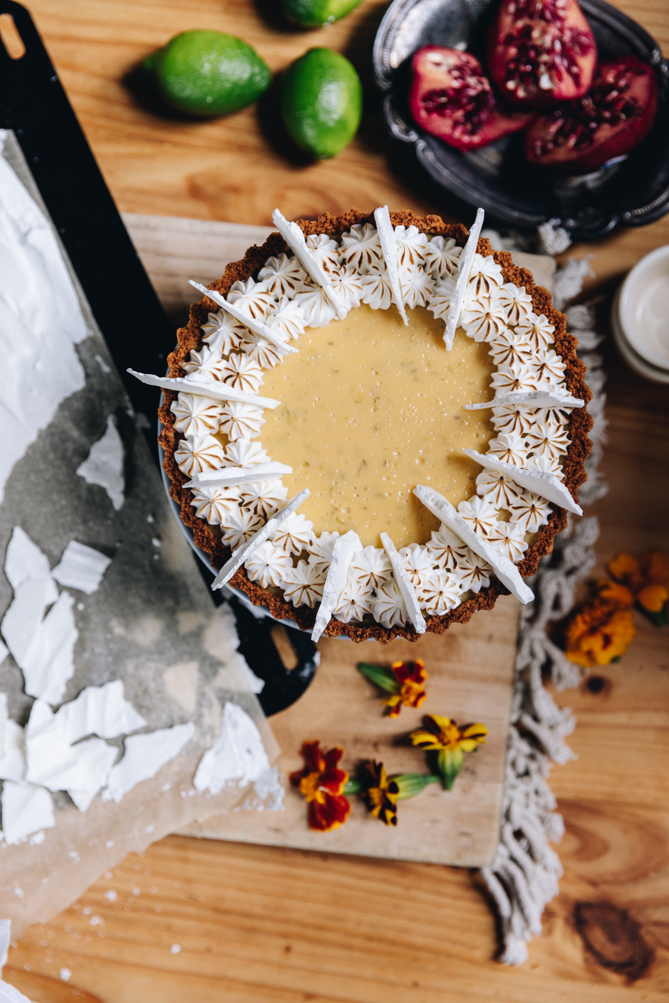 A flat lay shot shows a freshly decorated key lime pie recipe that has been decorated with piped meringue and meringue shards. The pie is on a wooden board and around the pie is limes, pomegranate pieces in a black small tray and some orange edible flowers. Next to the pie is a black tray with baking paper on it that has broken meringue shards on it.