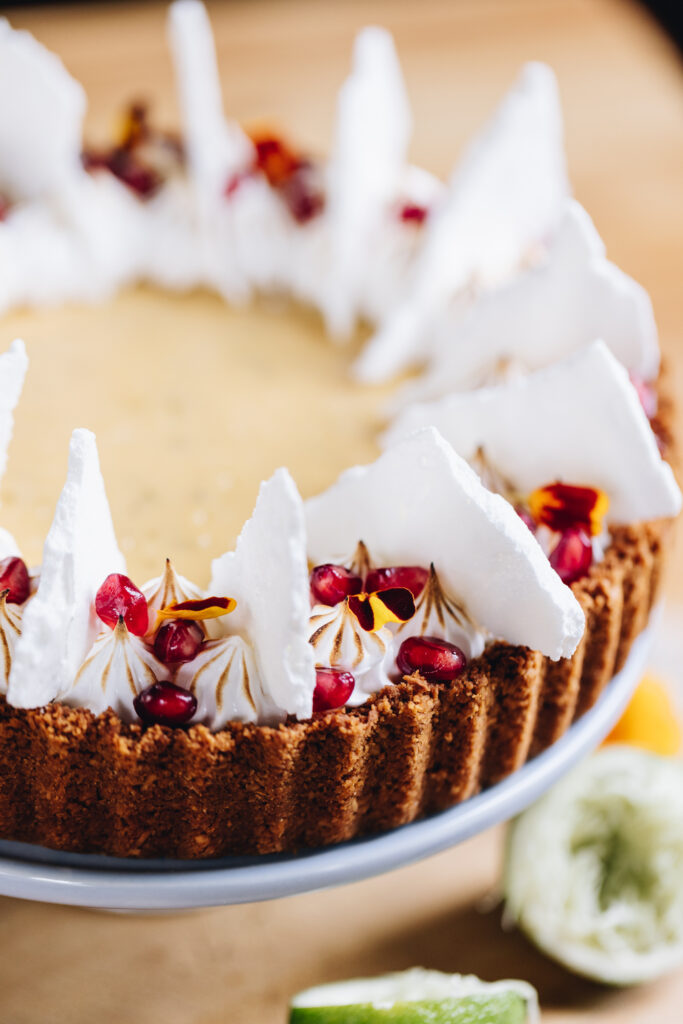 S freshly decorated key lime pie recipe has been decorated with piped meringue, crispy meringue shards, pomegranate seeds and flower petals. The pie is on a wooden board, sitting on a blue cake stand. Around the pie is cut limes.