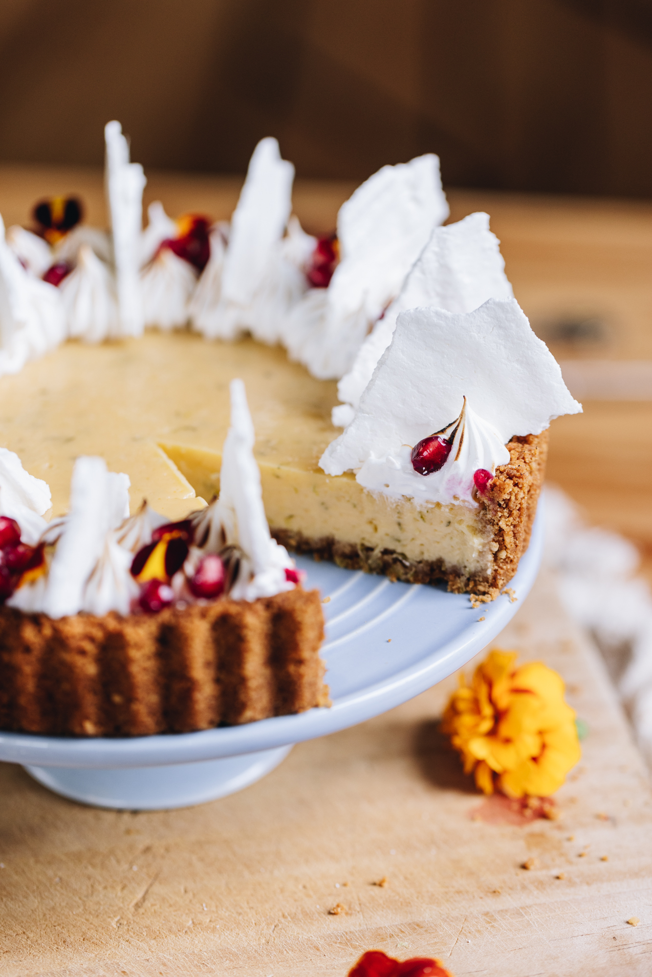 A freshly decorated key lime pie recipe has been decorated with piped meringue, crispy meringue shards, pomegranate seeds and flower petals. The pie is on a wooden board, sitting on a blue cake stand. A slice has been cut out of the pie, revealing the inside. A small orange edible flower is on the board.