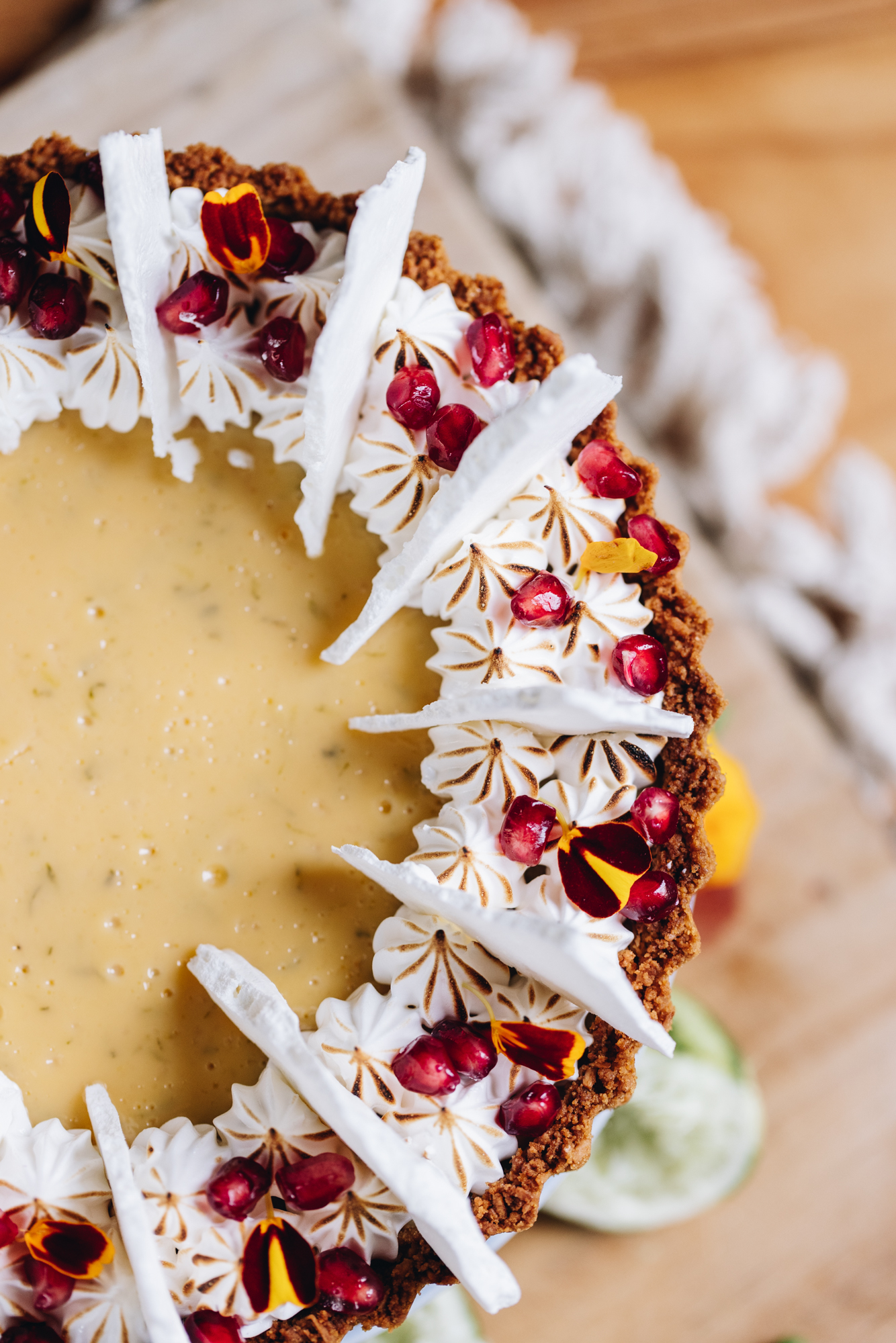 A flat lay shot shows a freshly decorated key lime pie recipe that has been decorated with piped French meringue recipe, meringue shards, pomegranate seeds and flower petals. The pie is on a wooden board that is sitting on a weaved cream mat.