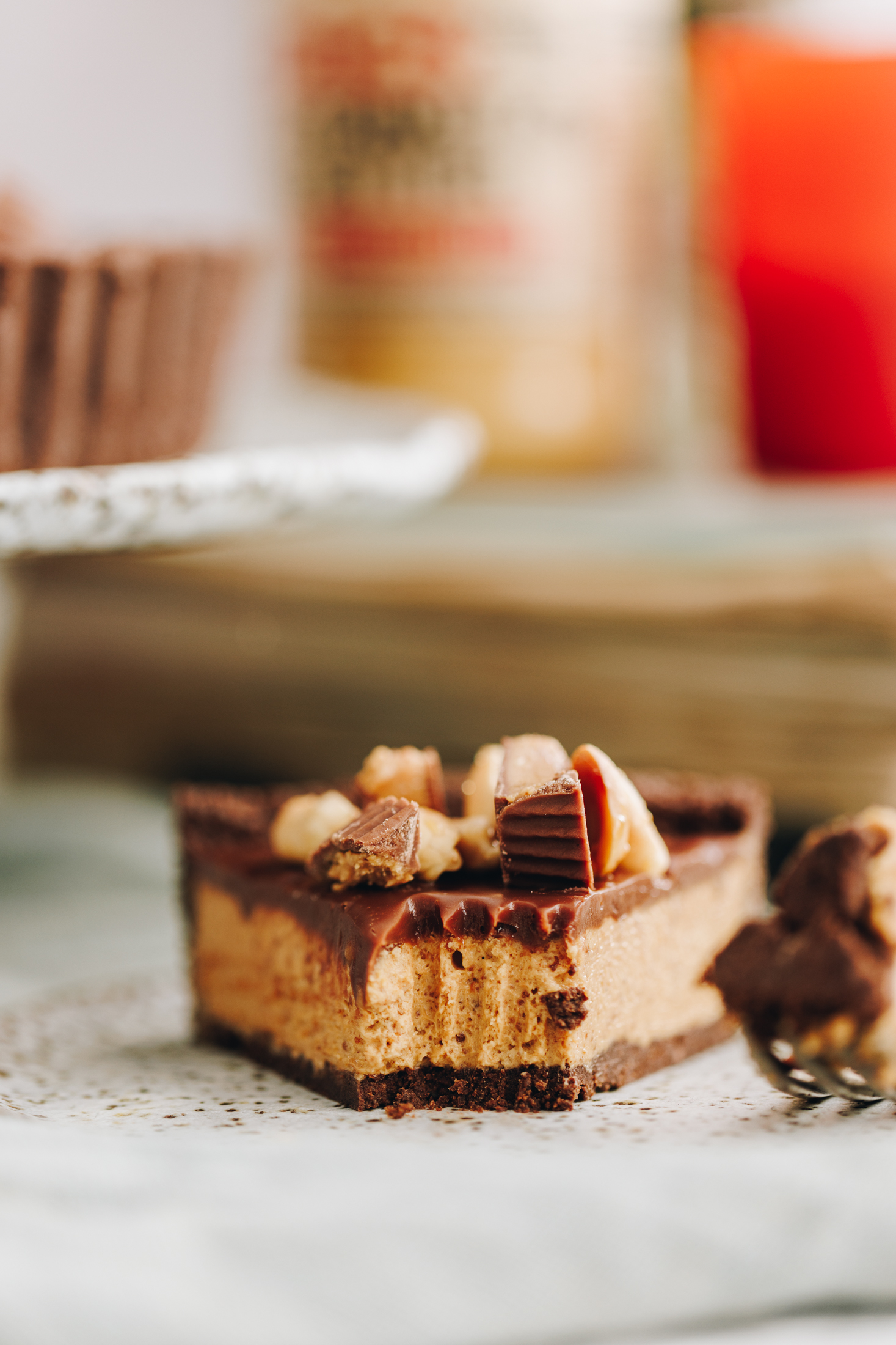 A slice of no-bake peanut butter pie sits on a grey table. It has a fork piece removed that is next to it and the chocolate biscuit base, the creamy peanut butter filling and the chocolate topping is seen. There are mini peanut butter cups and sticky peanuts on top too. In the background is a cake stand with the pie blurred behind it. A pics peanut butter jar and an orange candle are also blurred in the background. 
