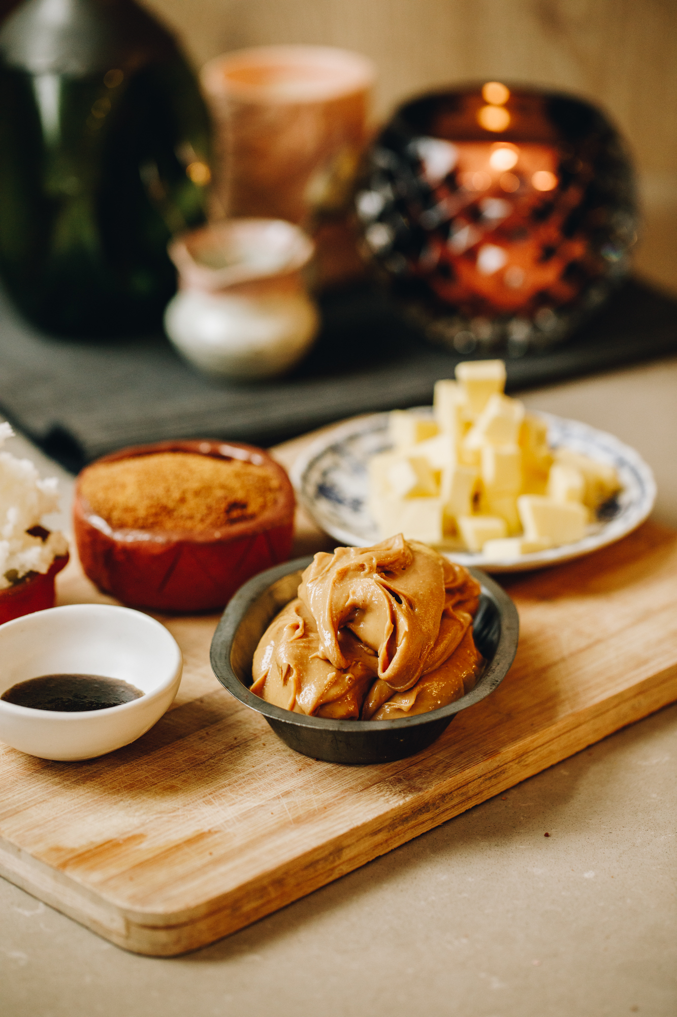 A wooden boards sits on a light brown bench. On the board is a small tray of peanut butter, a small bowl of vanilla, a ceramic small bowl has coconut sugar and a plate has cubed butter on it. In the background is a black glass candle holder with a candle in it. There is also a small jug, a green glass jar and an orange candle in the background sitting on a black mat. 