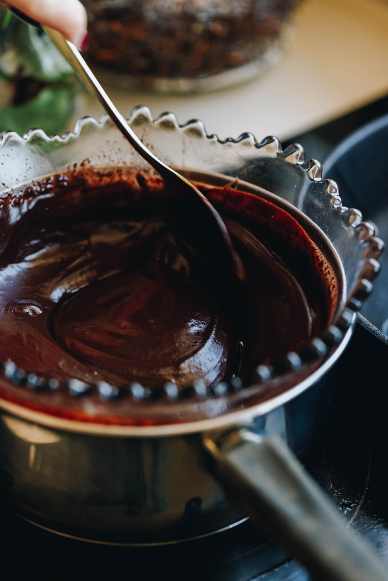 A purple vintage bowl sits snug in a silver pot on top of a black stove. In the bowl is melted chocolate and Naomi is stirring it with a spoon. 