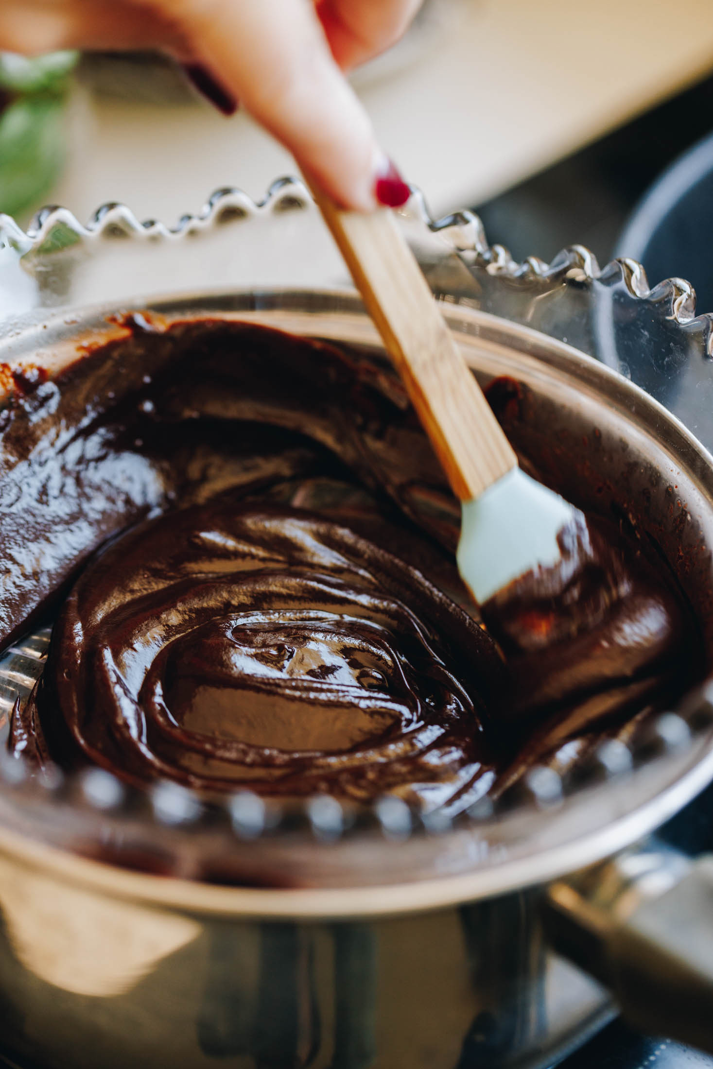 A close up shot shows a purple vintage bowl sitting snug in a silver pot on top of a black stove. In the bowl is melted chocolate and Naomi is stirring it with a spoon.
