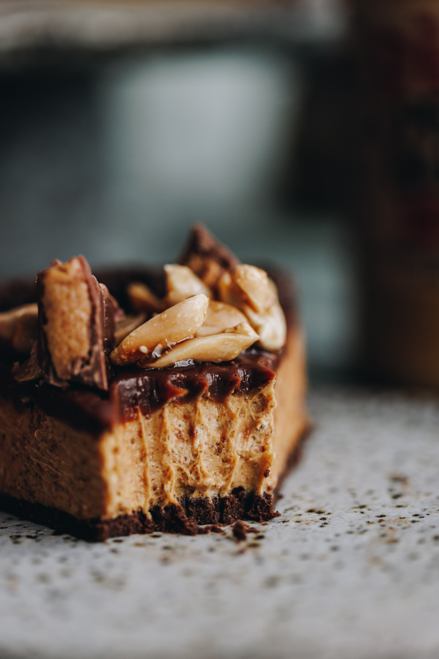 A super close slice of no-bake peanut butter slice shows the sits on a grey plate. It has a fork piece removed that is next to it and the chocolate biscuit base, the creamy peanut butter filling and the chocolate topping is seen. There are mini peanut butter cups and sticky peanuts on top too. 
