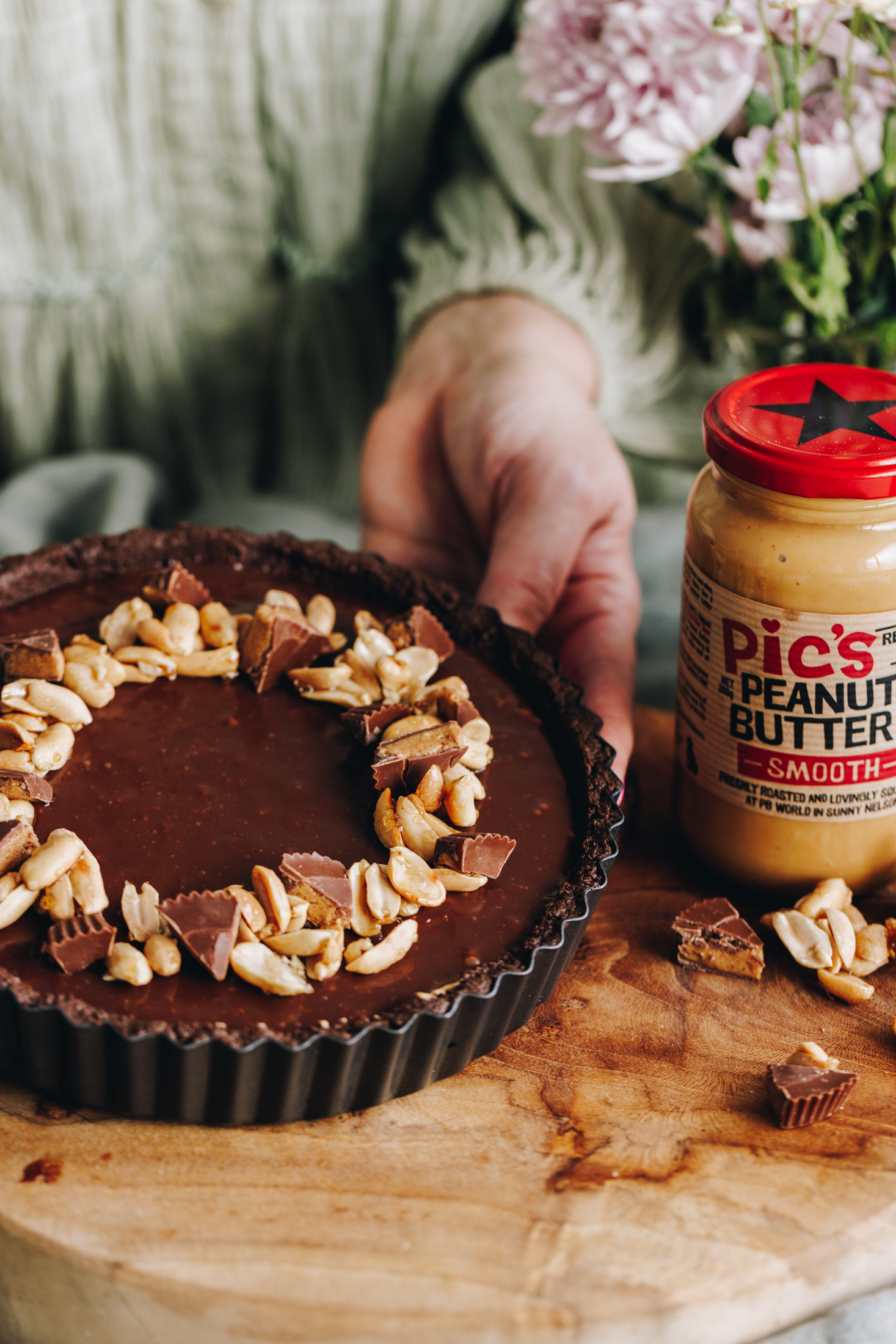 A no-bake peanut butter pie is in a quiche tin and is being tipped forward on a wooden board. There is chocolate ganache on top and maple peanuts are arranged in a circle in the centre. Mini peanut butter cups are cut in to pieces and are arranged in between the nuts. A smooth Pic's peanut butter jar is next to it with pink flowers behind it.