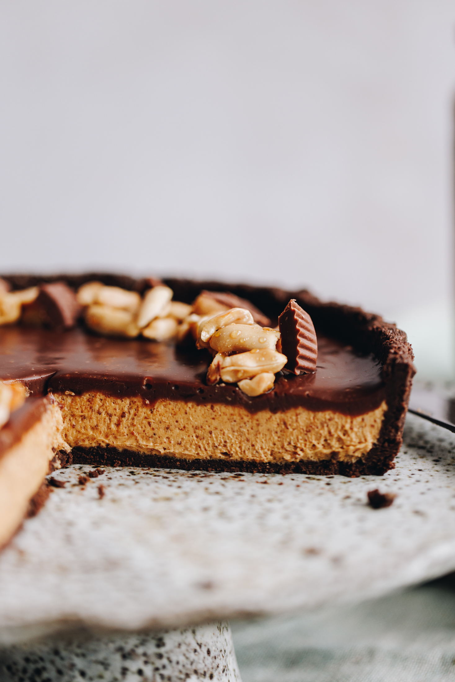 A no-bake peanut butter pie is on a ceramic cake stand and has been cut open to reveal the chocolate biscuit base, the creamy peanut butter filling and the chocolate topping. There are mini peanut butter cups and sticky peanuts on top too. 
