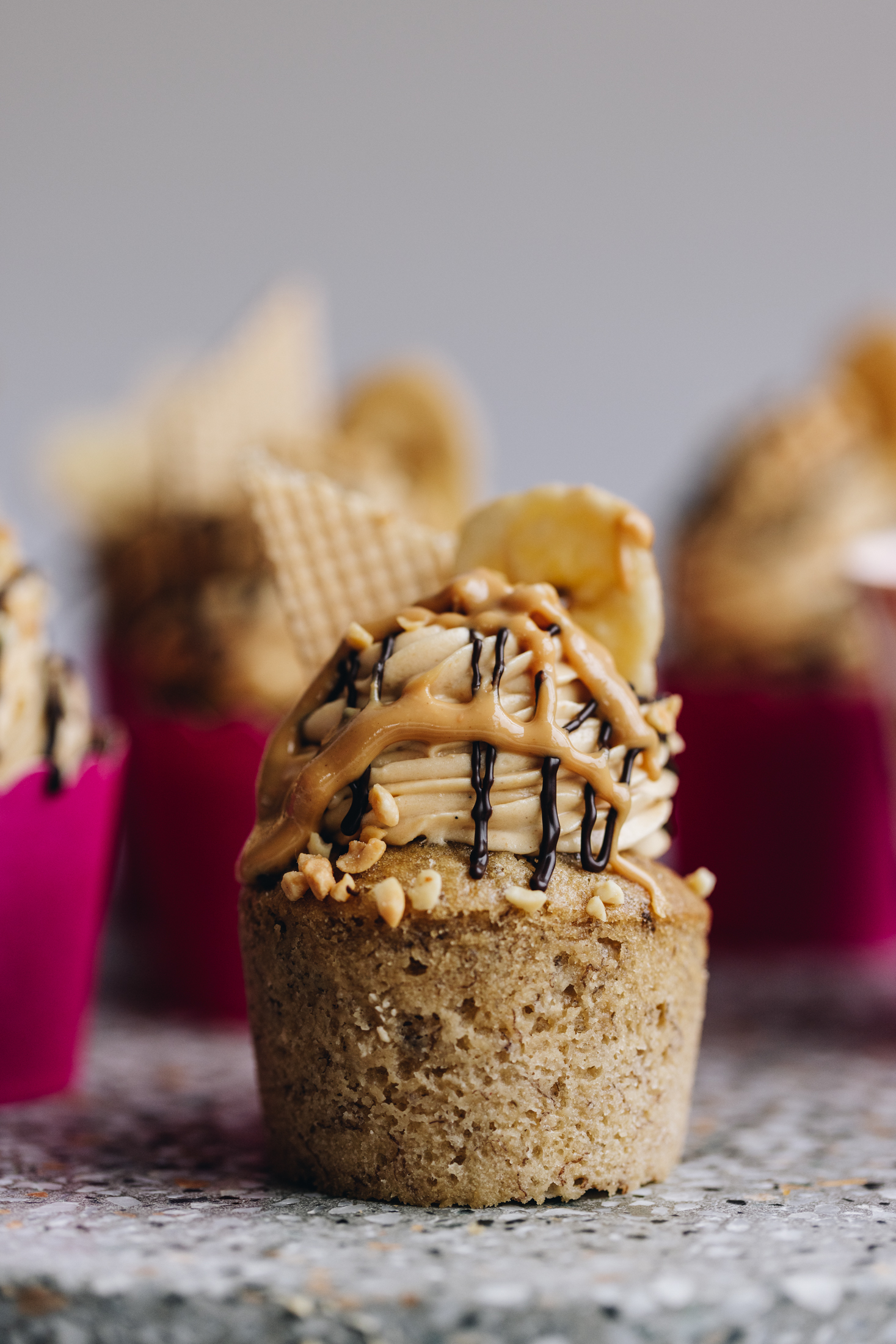 Simple banana cupcakes sit on grey stone. One has had the pink cardboard removed and shows the cupcake with peanut butter frosting, a slice of wafer, a banana chip and a drizzle of dark chocolate on top. There is other cupcakes around this one that are in pink cases, blurred in the background.