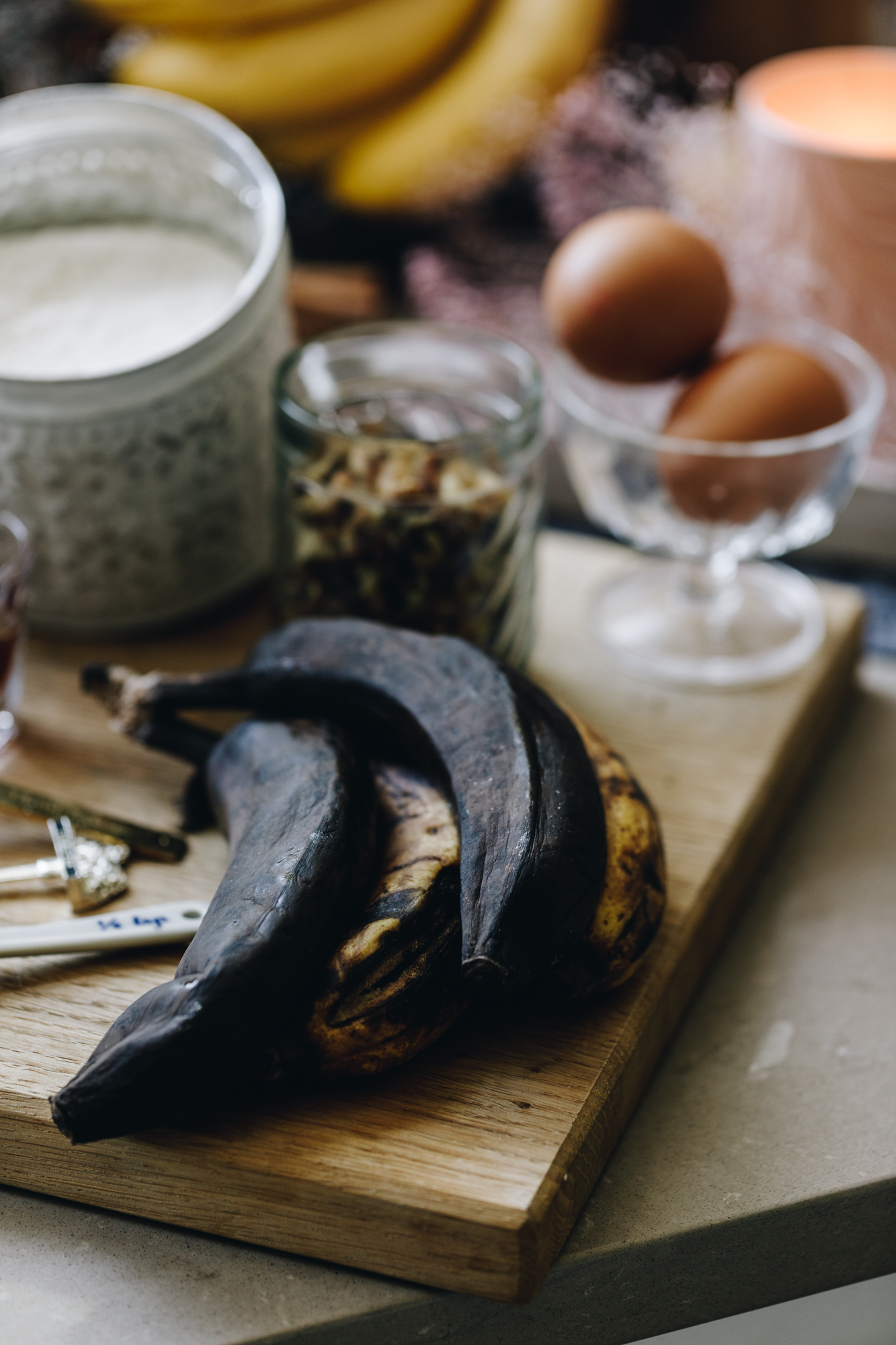 A wooden board sits on a brown bench. On the board is four rotten banana, some vintage teaspoons, a glass jar with two eggs in it, a glass with nuts and a white metal jar with flour. Behind the board is an orange burning candle and a bunch of yellow bananas.
