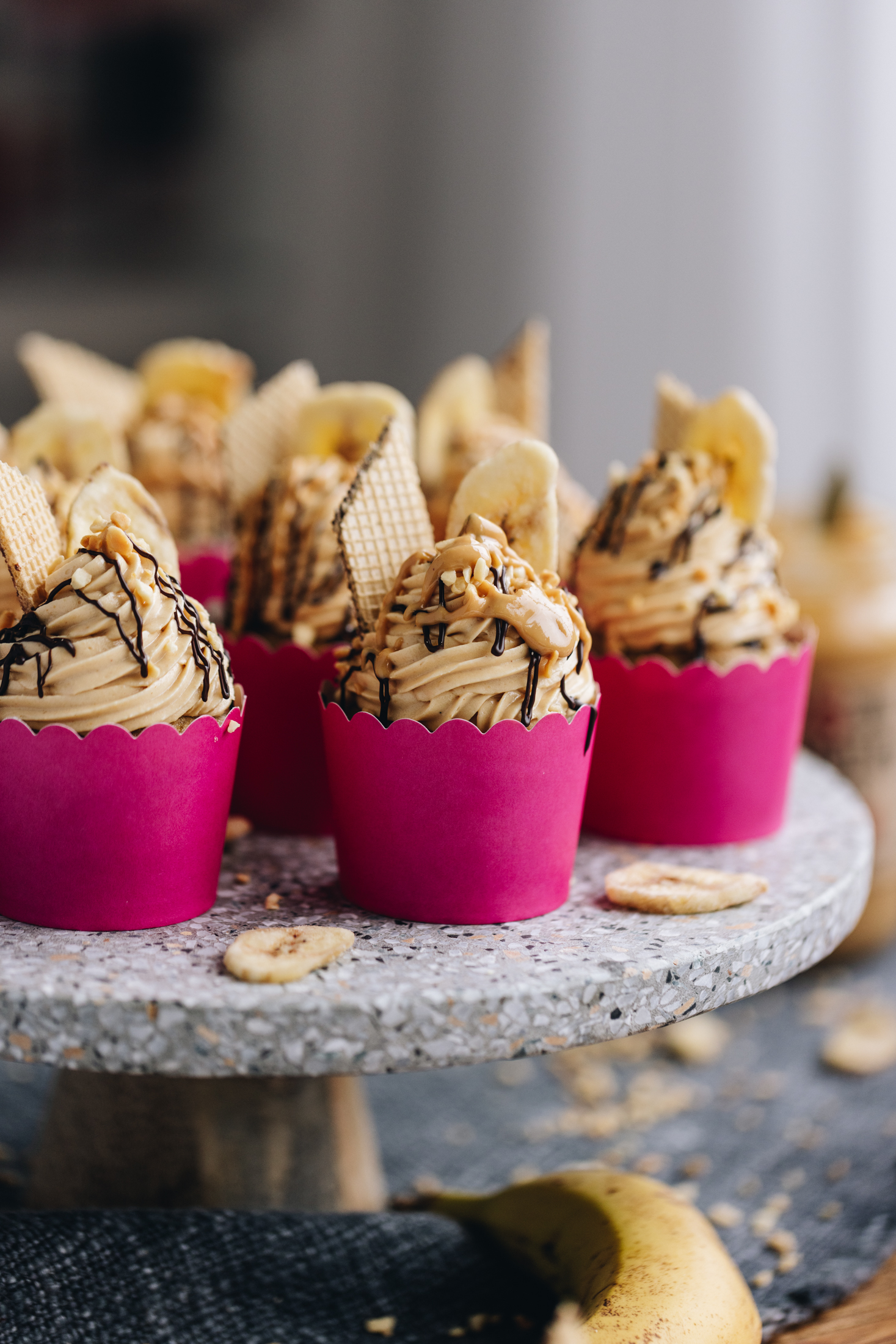 A speckled grey stone cake stand is on a table with a dark cloth under it. On the stand is freshly decorated simple banana cupcakes decorated with peanut butter frosting, wafers, banana chips and a drizzle of dark chocolate on top. There are other cupcakes around this one that are in pink cases, blurred in the background. Banana chips are next to the cupcakes and a banana is at the forefront of the shot.