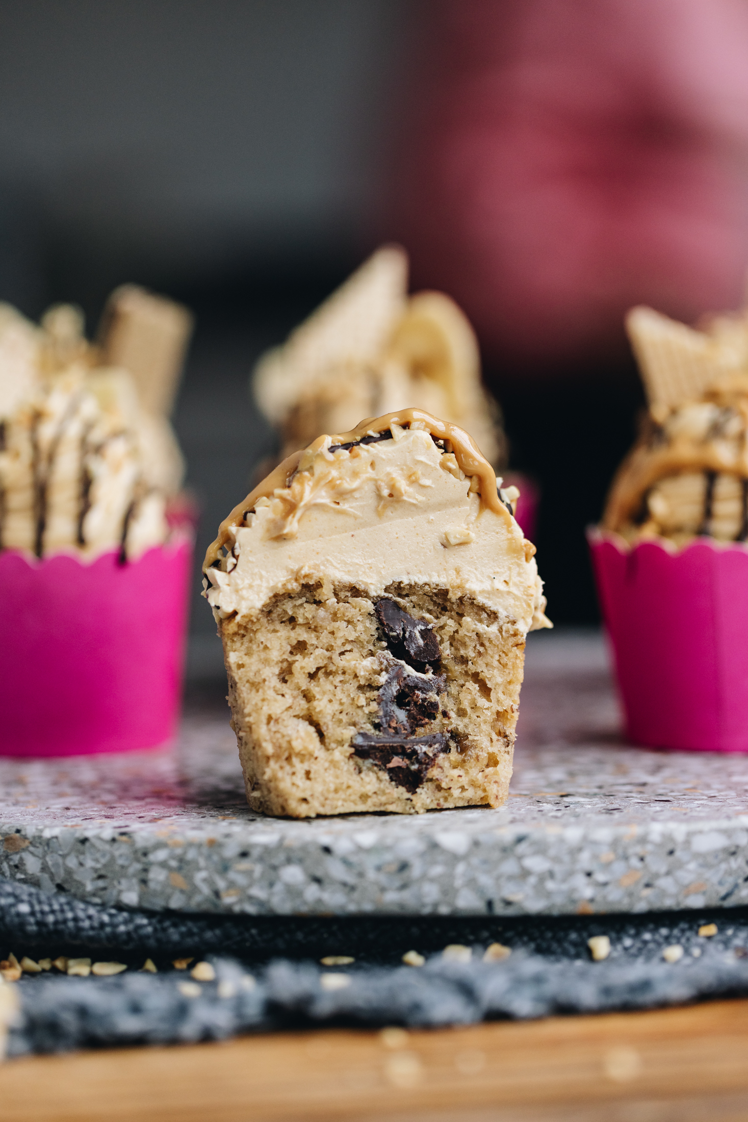 A simple banana cupcake is on grey speckled stone. It has had the paper removed and has been cut open to reveal the chocolate hidden inside the cupcake. It has been topped with peanut butter buttercream and a touch of peanut butter. There are other cupcakes around this one that are in pink cases, blurred in the background.