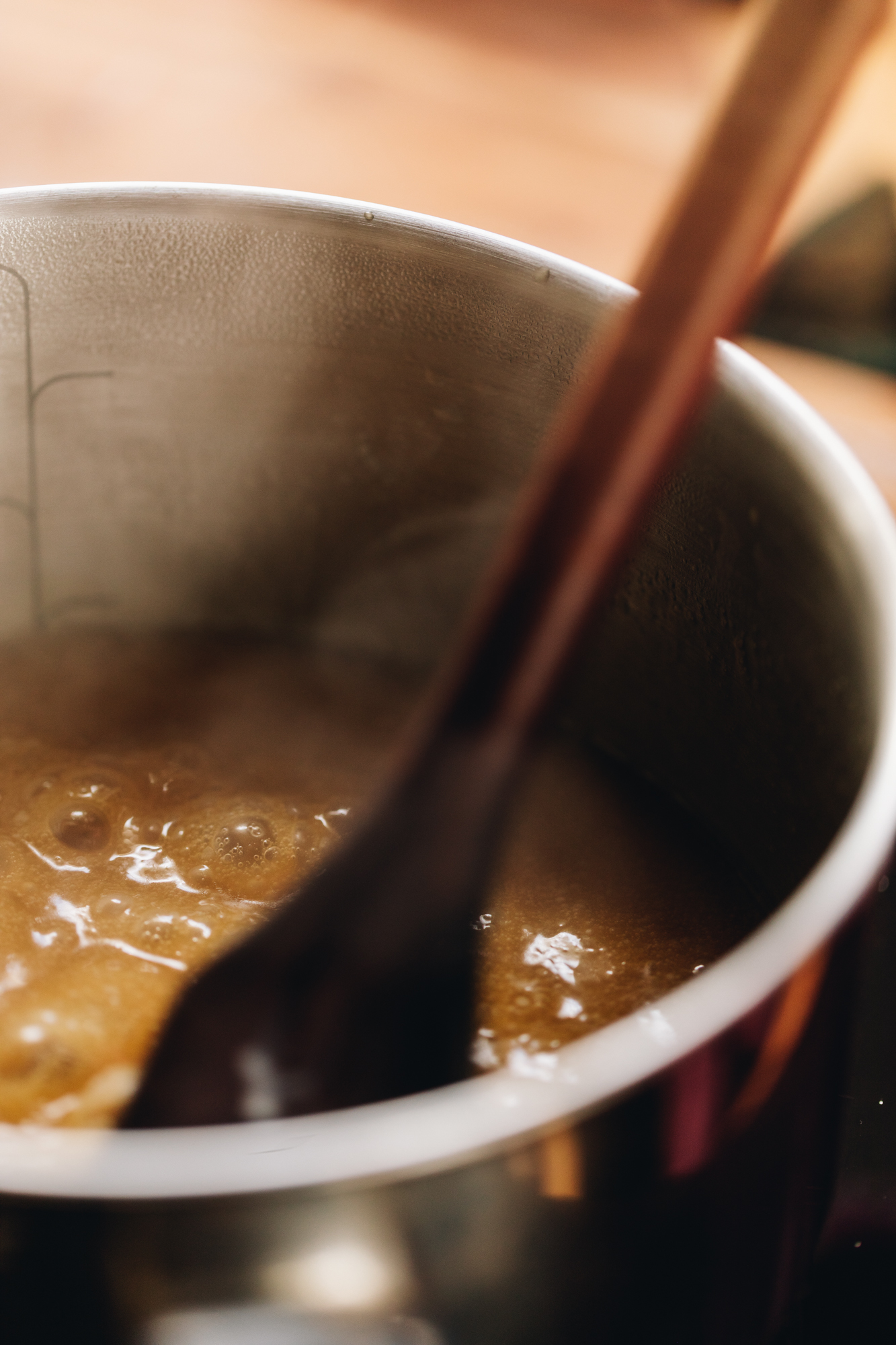 In a silver pot is a thick caramel that is boiling. A wooden spoon is stirring it at the same time.