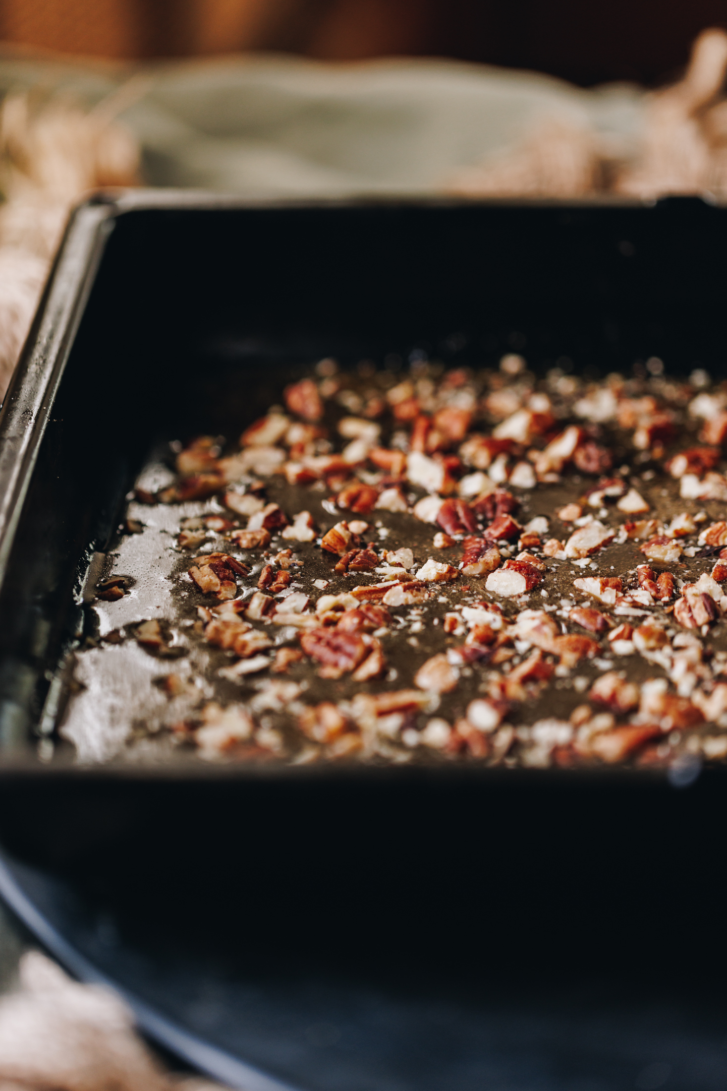 A black tray is sitting on a table limed with a green linen tablecloth. In the tray is gooey maple syrup  caramel with chopped pecan nuts on top. 