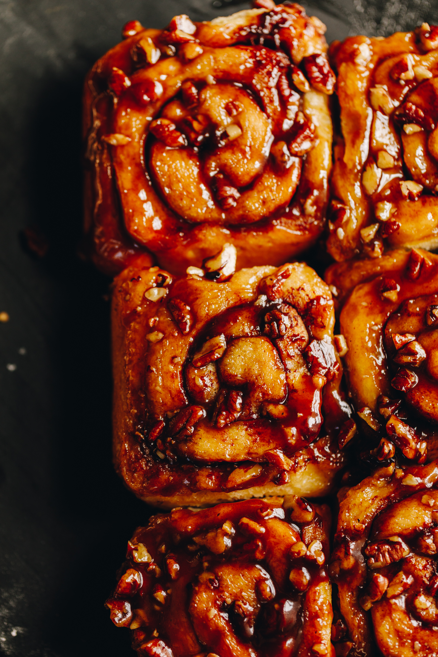 Freshly baked sticky maple cinnamon scrolls have been flipped over. The flat lay shot reveal the sticky maple caramel and toasted pecans that are glossy on top of the scrolls. They are on a black tray. 