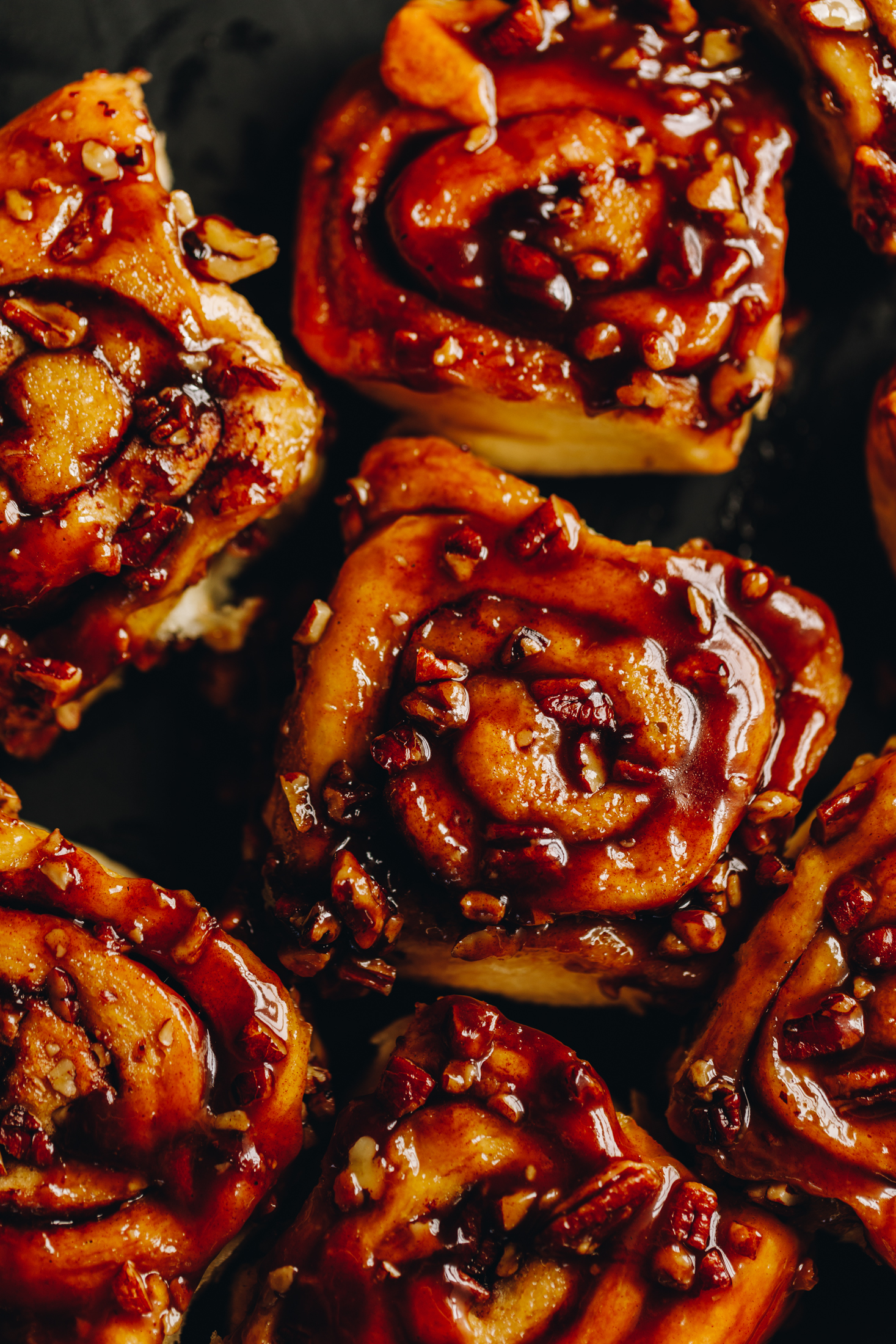 Freshly baked sticky maple cinnamon scrolls have been flipped over and have been pulled apart. The flat lay shot reveal the sticky maple caramel and toasted pecans that are glossy on top of the scrolls. They are on a black tray.