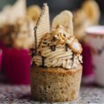 A single banana cupcake has been unwrapped and shows the banana cupcake texture, the peanut butter buttercream on top with extra peanut butter, chocolate drizzle, a wafer and banana chip on top.