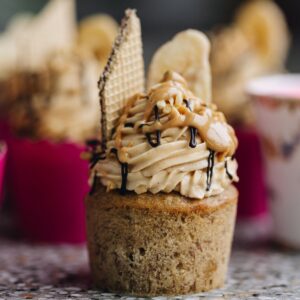 A single banana cupcake has been unwrapped and shows the banana cupcake texture, the peanut butter buttercream on top with extra peanut butter, chocolate drizzle, a wafer and banana chip on top.