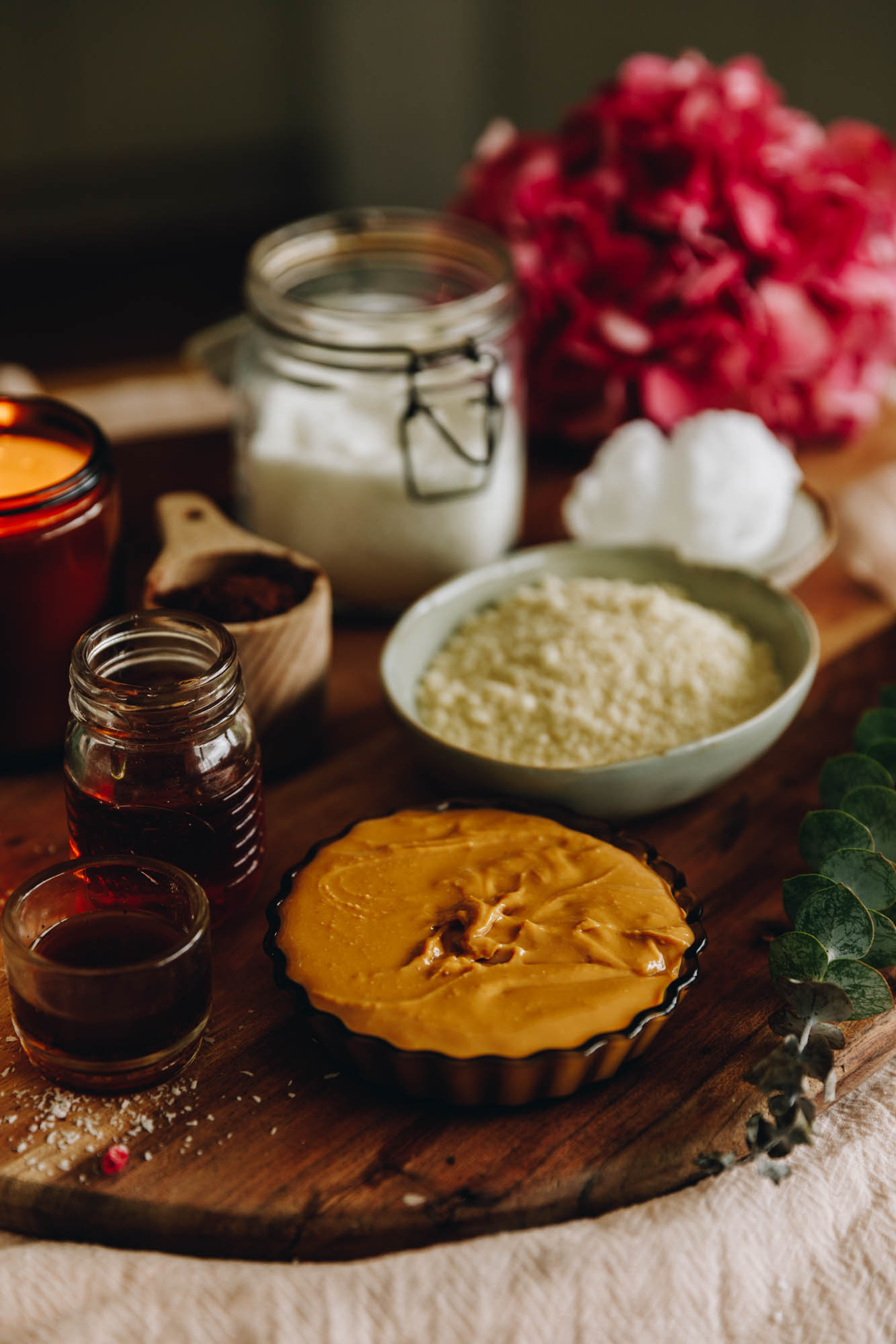 On a wooden board sits vintage bowls and small plates that have pan cotta ingredients on it. There is peanut butter, biscuits and vanilla in view. They are sitting on a wooden table with natural fabrics on it with pink flowers are in the background.
