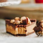 A slice no-bake peanut butter pie is on a speckled plate, fork marks are seen through the front of the slice revealing the inside. Chocolate biscuit base with peanut butter filling, chocolate top and mini peanut butter cups.