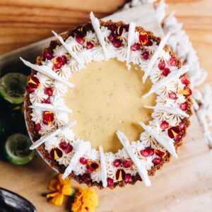 A flat lay of decorated and baked key lime pie that has the key lime custard in the centre with piped meringue around the edge with pomegranate seeds and flower petals and meringue shards on top.